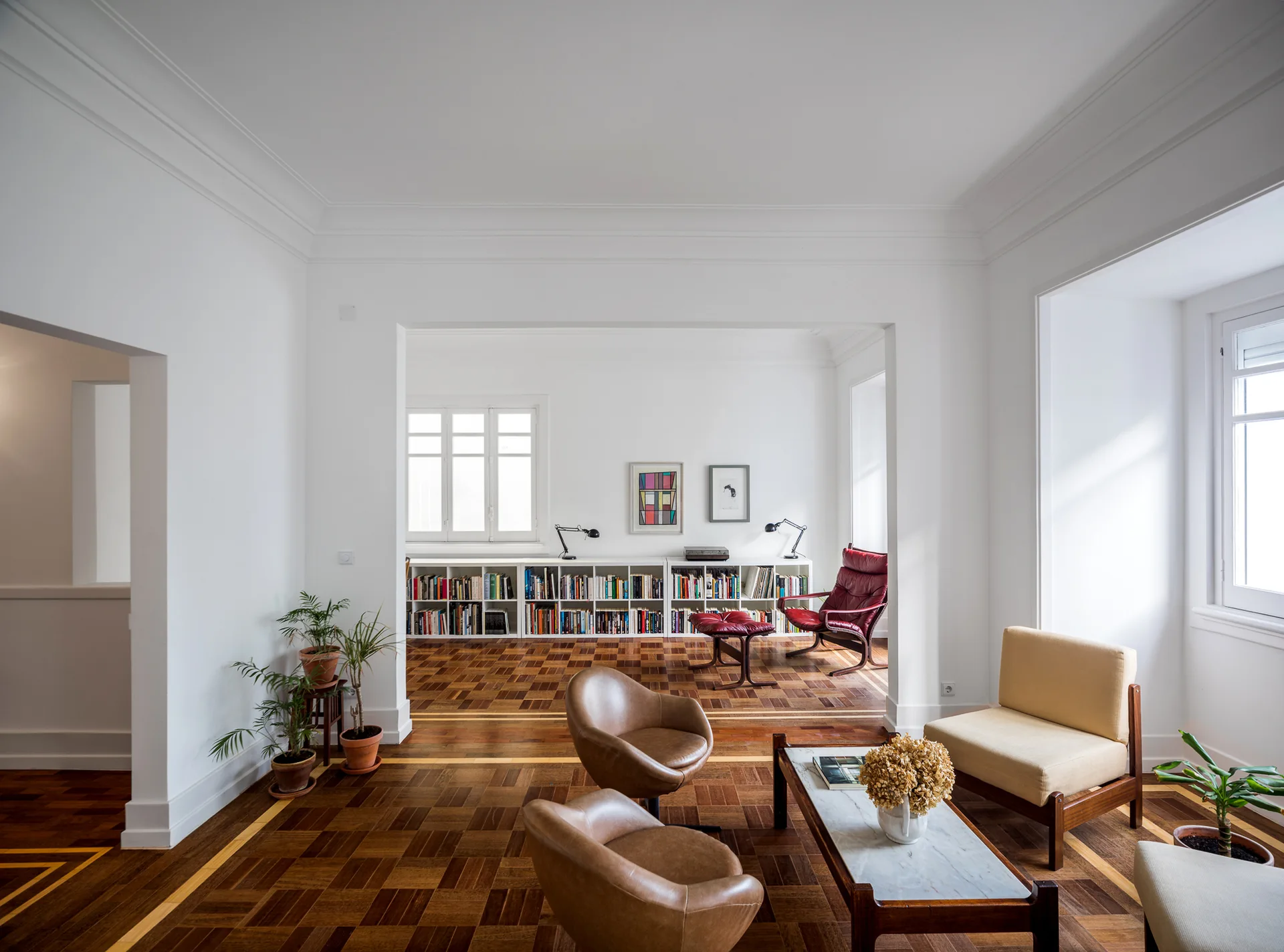 An open plan living room features white walls, geometric parquet wood flooring, mid century leather armchairs, and a low white bookcase.