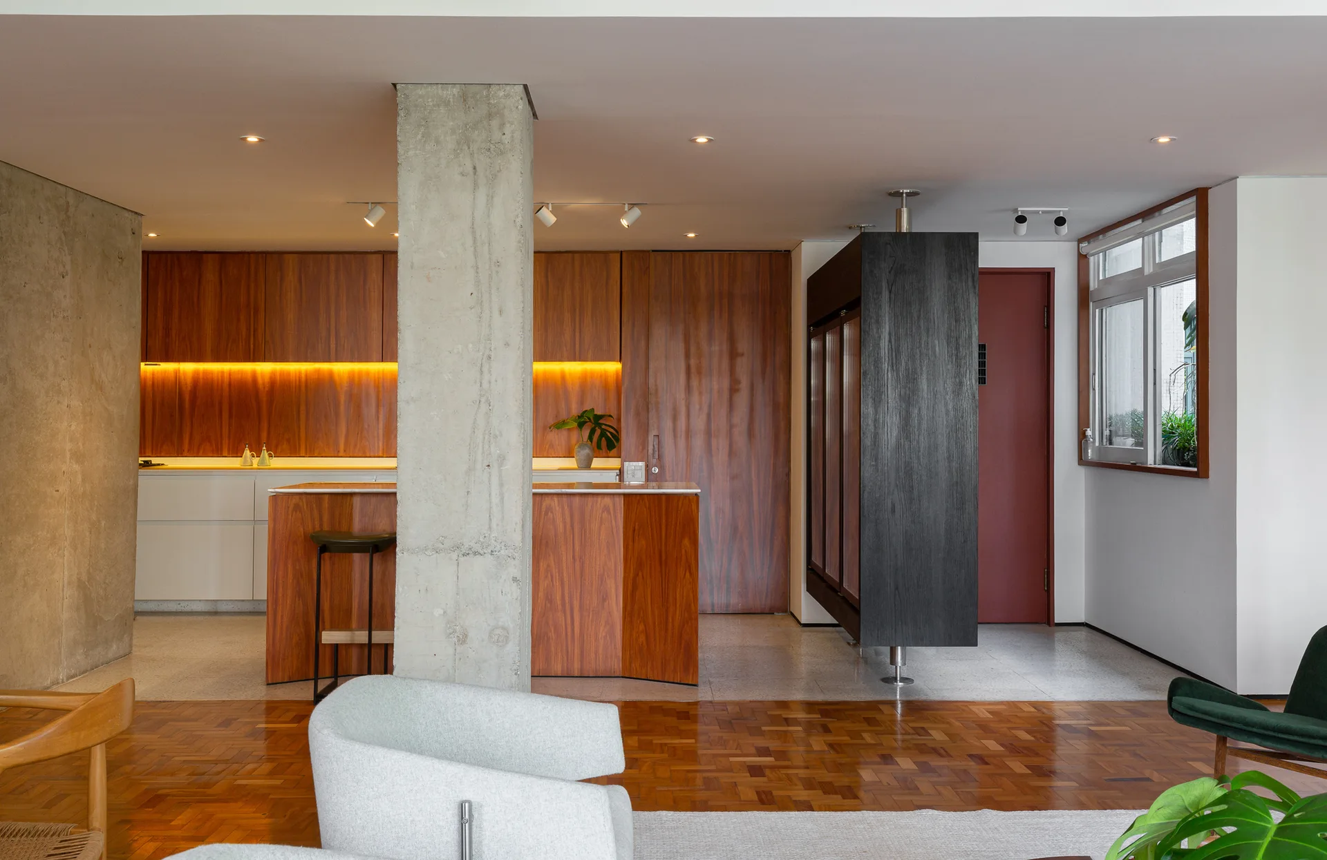 A modern open-plan kitchen and living space featuring warm wood cabinetry, a raw concrete structural column, and parquet flooring.