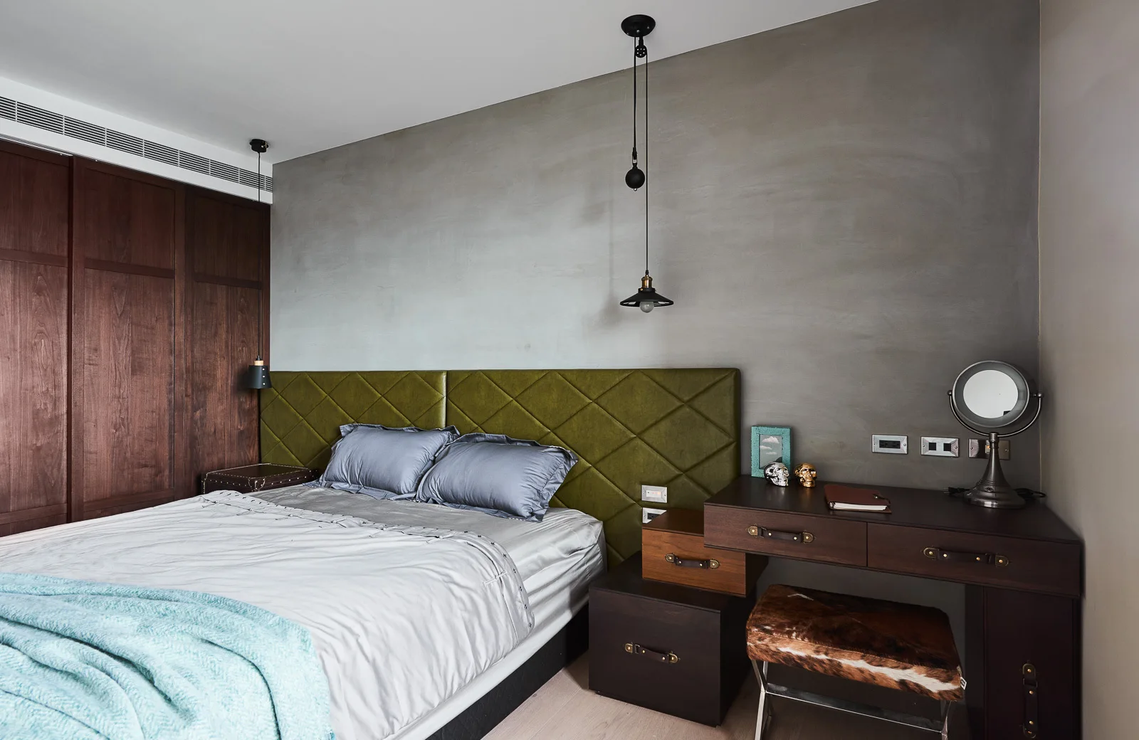 A bedroom features a bed with a quilted olive green headboard set against a textured gray wall beside a dark wood vanity desk.