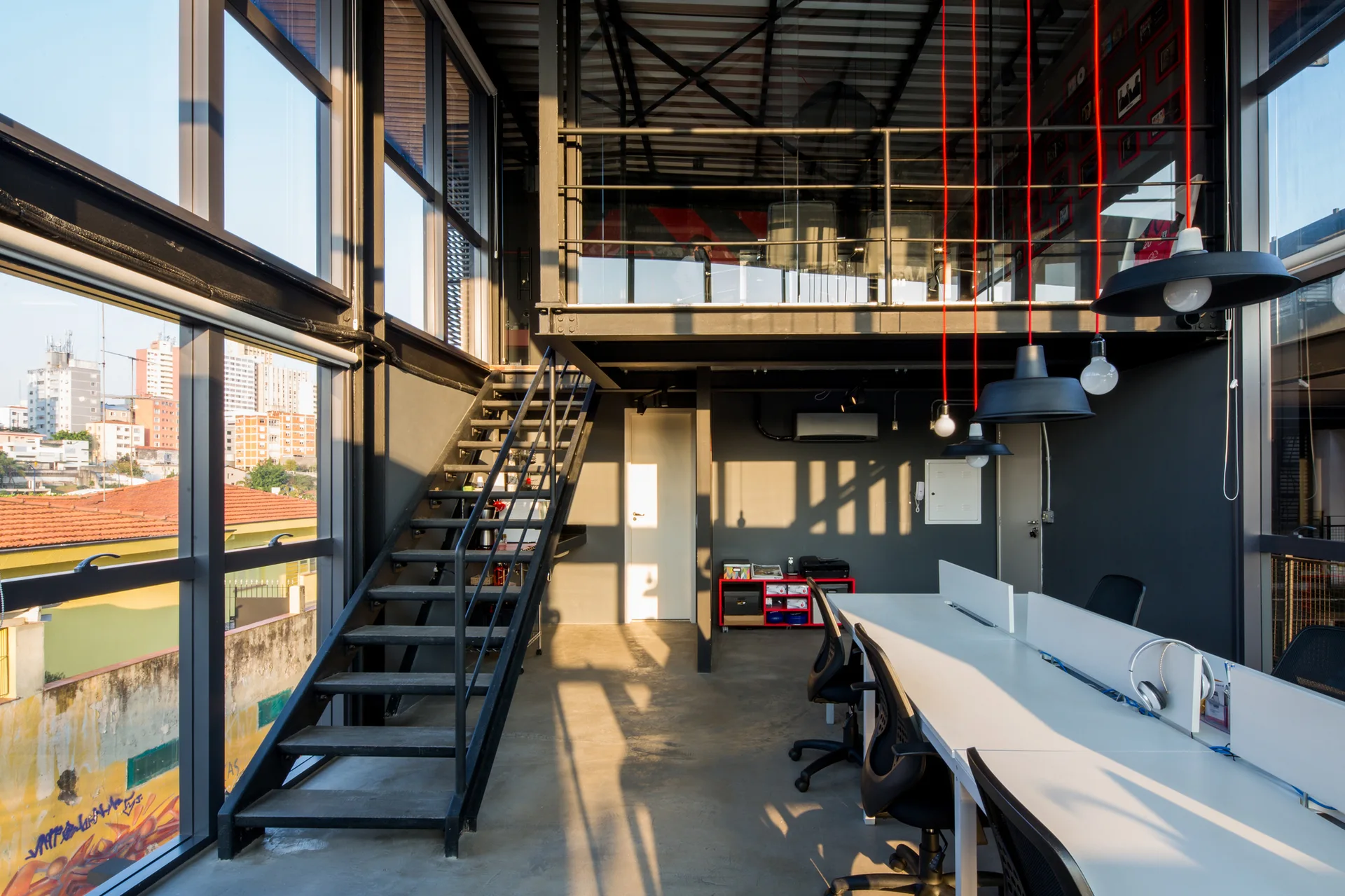 An industrial office featuring a long white desk with task chairs, a black metal staircase leading to a mezzanine, and large windows.