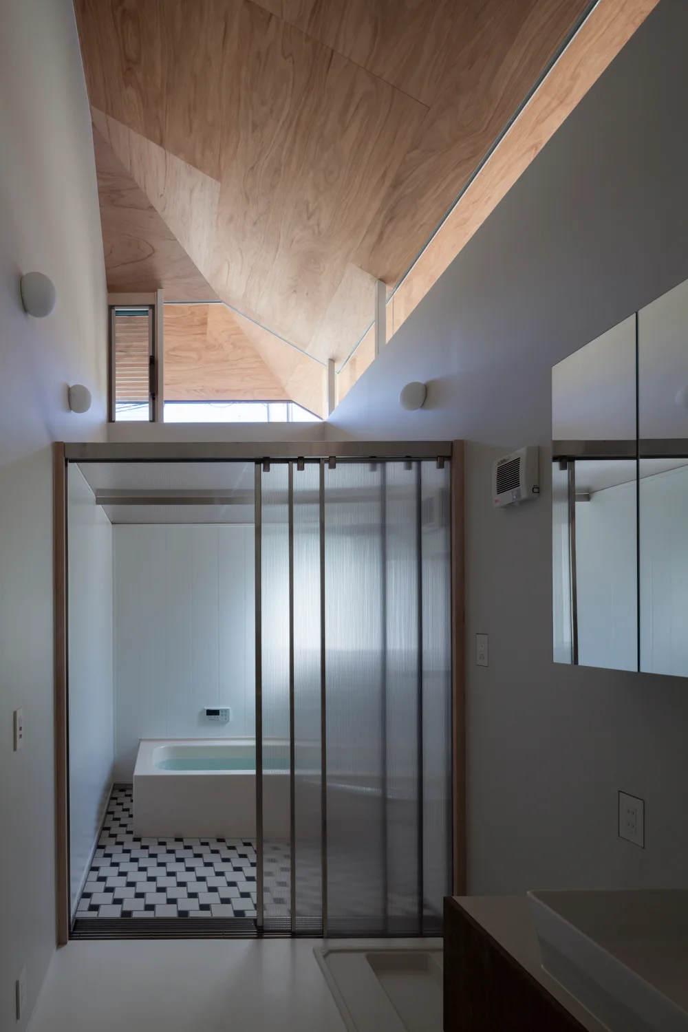 A minimalist bathroom features an angled wood-paneled ceiling, a fluted glass sliding door, and black-and-white checkered floor tiles around a white bathtub.