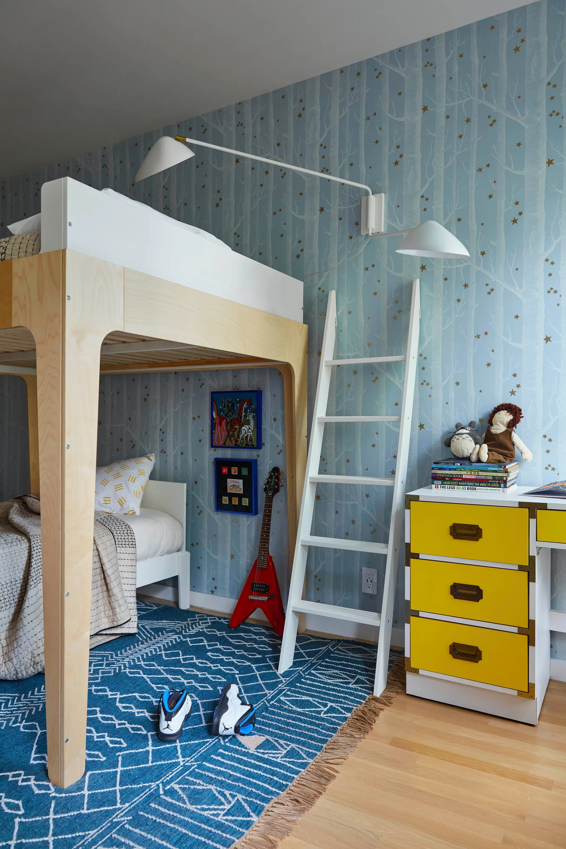 A children's bedroom features a light wood bunk bed, a blue forest-patterned wallpaper, a white ladder, a blue geometric rug, and a yellow drawer unit.