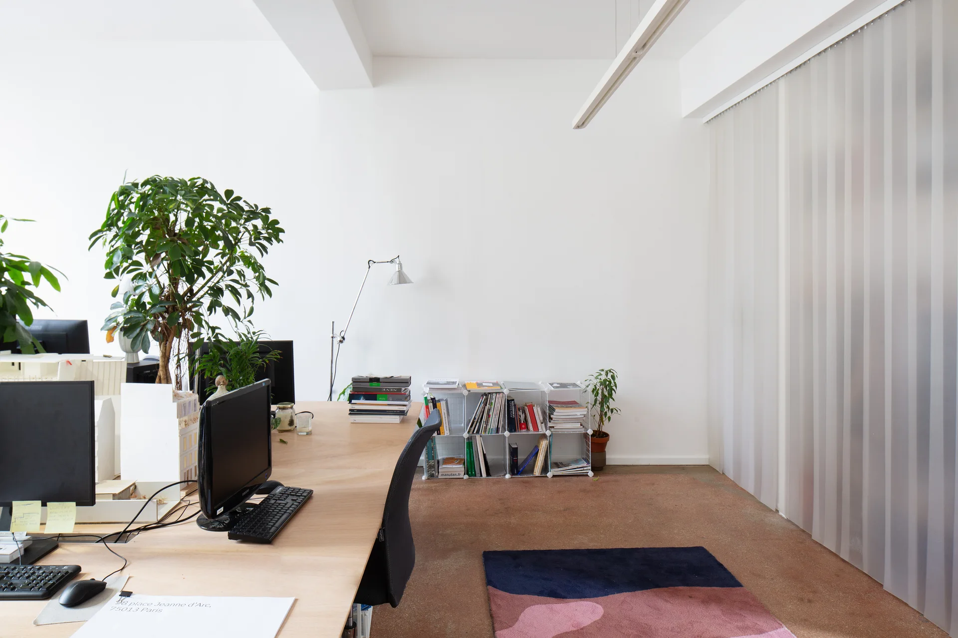 A minimalist office featuring a long wood desk with computer monitors, a low wire bookshelf on a concrete floor, and translucent vertical blinds.