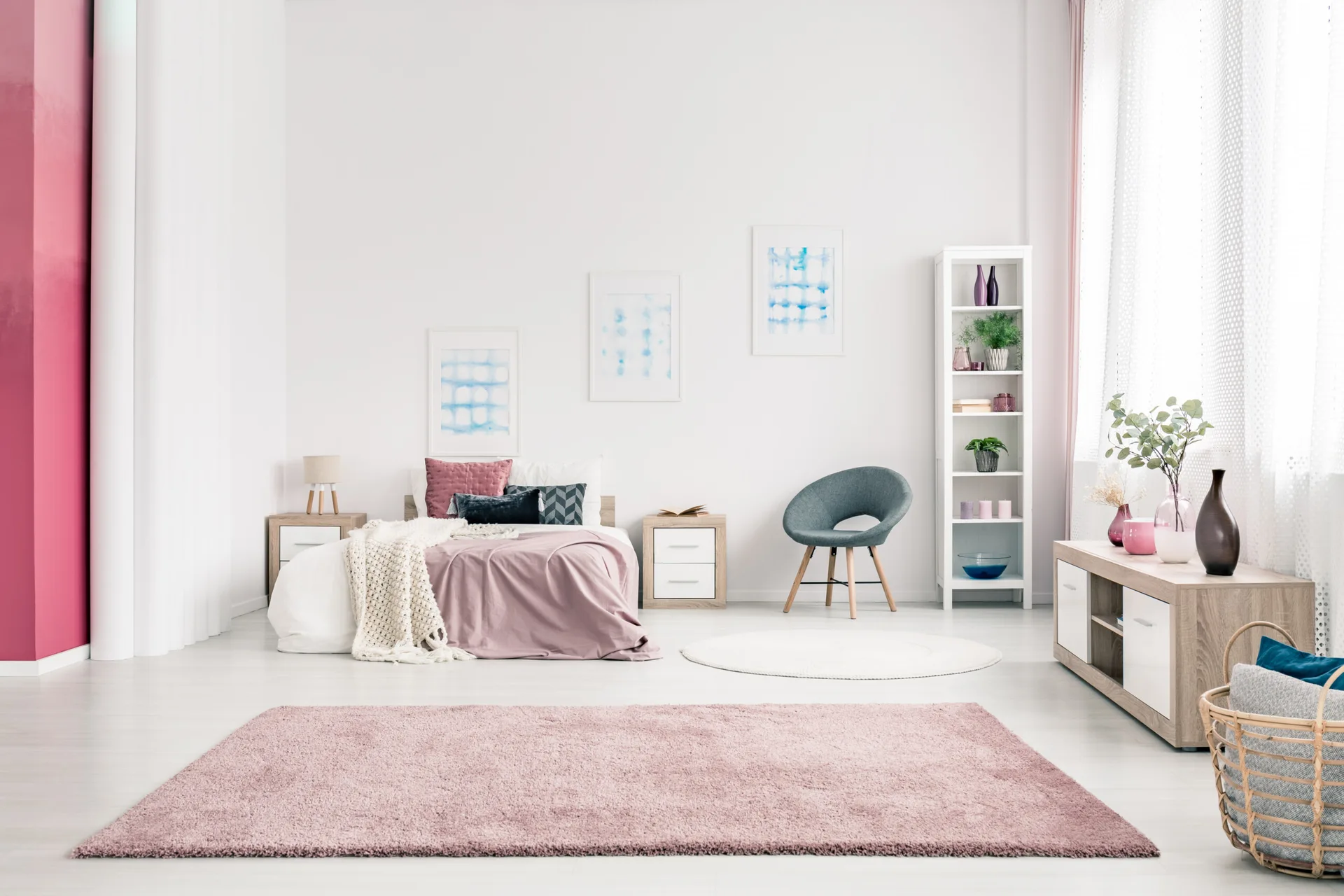 A bright bedroom features a bed with pink bedding, two light wood nightstands, a pink area rug, and a tall white shelving unit.