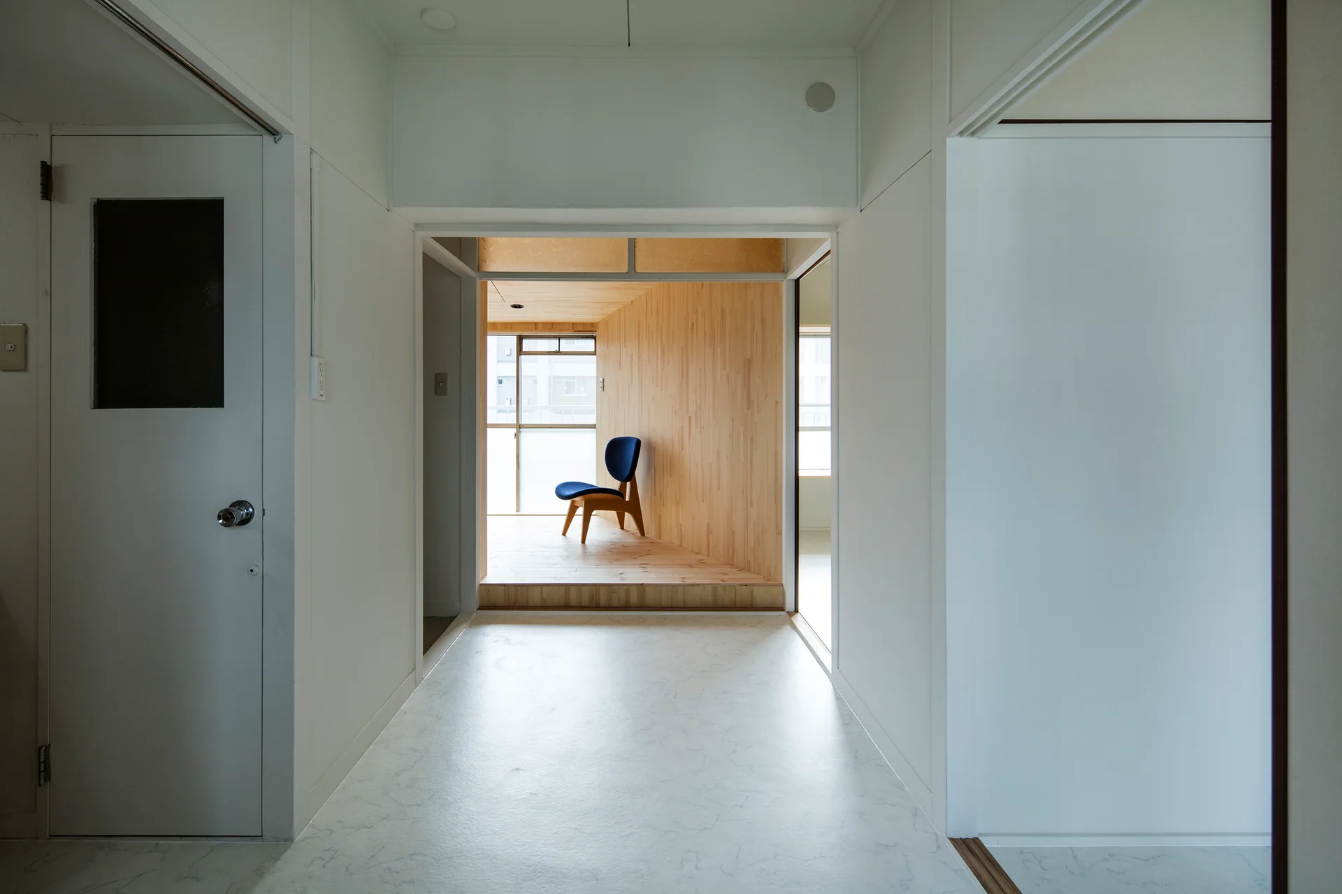 A minimalist white hallway leads to an elevated, wood-paneled room featuring a single dark blue chair in front of a window.