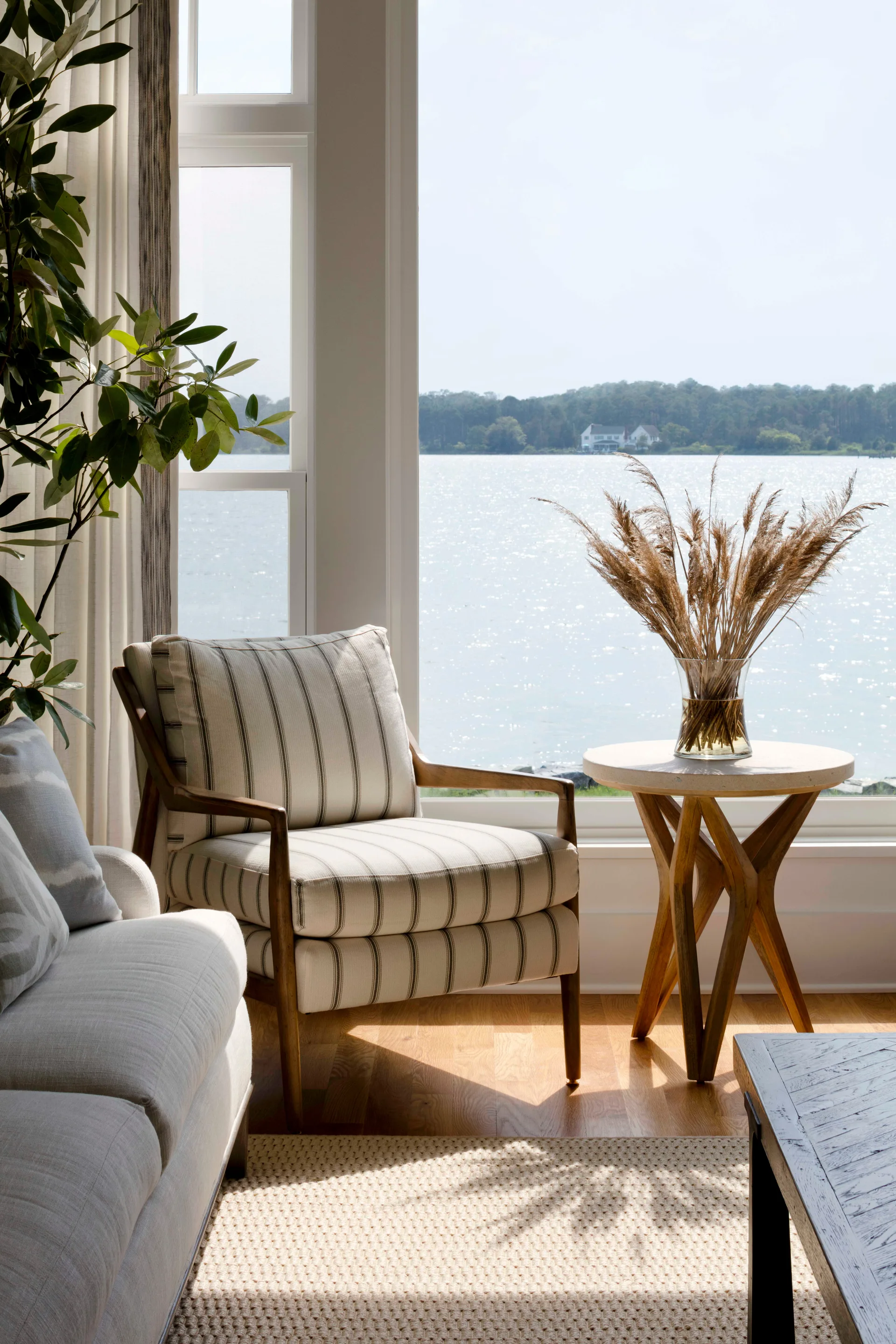 A striped upholstered armchair sits next to a wooden side table with dried grasses in a glass vase, framed by a window overlooking a quiet lake.