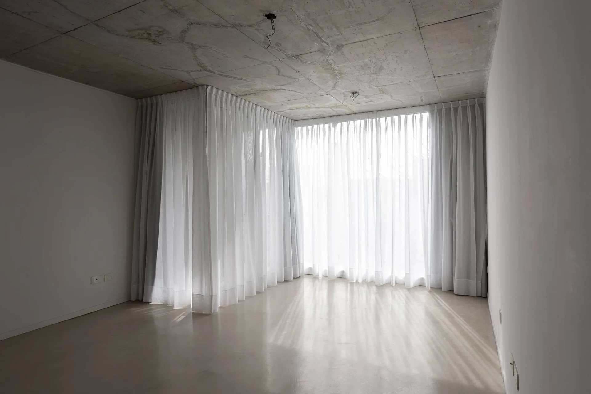 An empty minimalist room featuring an exposed concrete ceiling, smooth light flooring, and sheer white floor-to-ceiling curtains covering corner windows.
