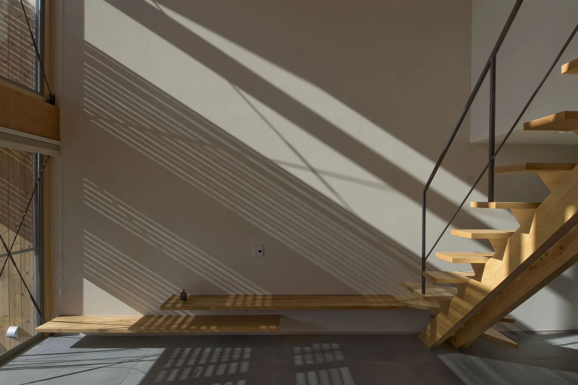 A light wood open riser staircase rises next to two matching low floating shelves on a beige wall cast with geometric window shadows.