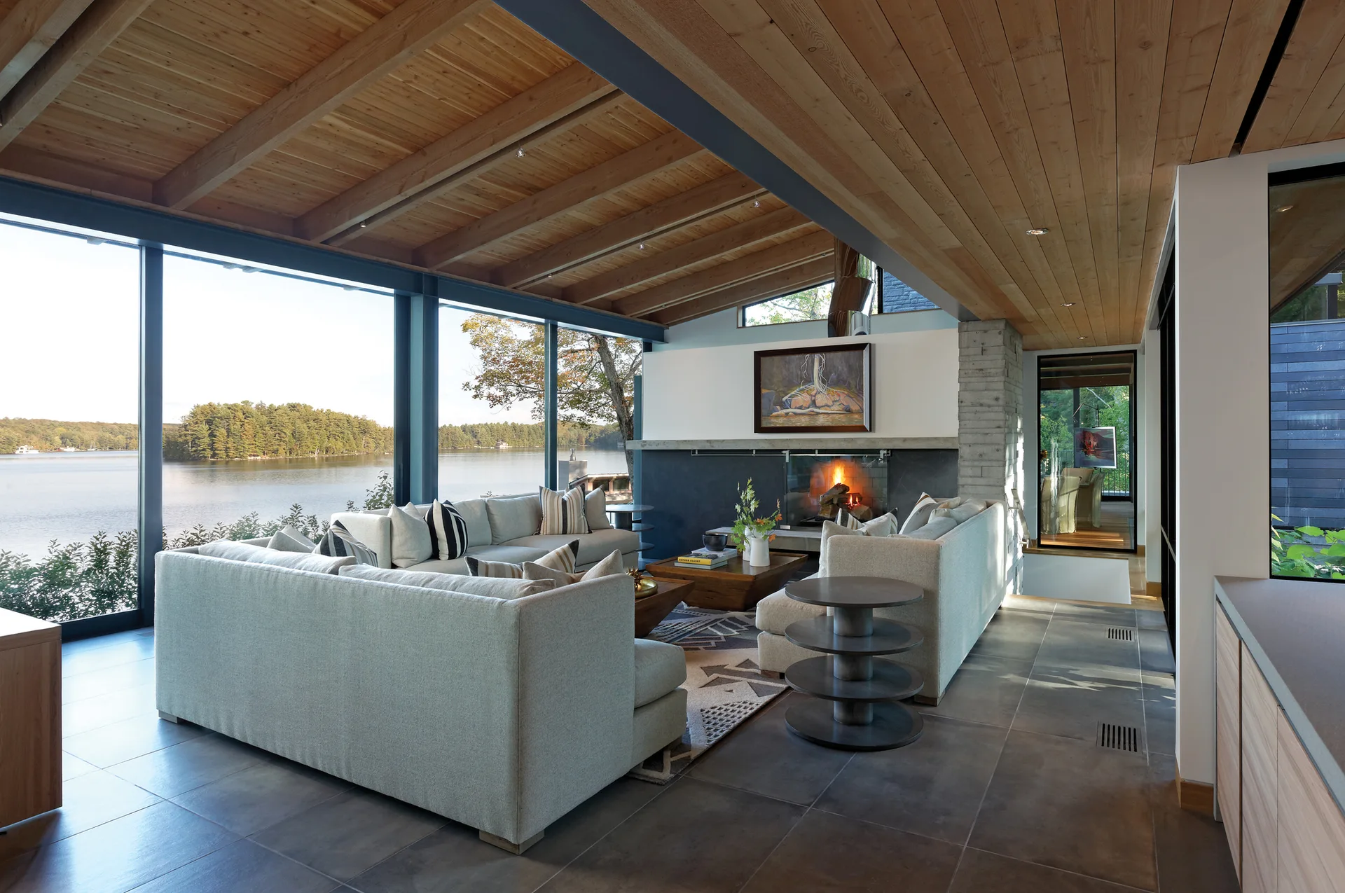 A modern lakeside living room features a light grey sectional sofa facing a fireplace under a sloped wood ceiling.