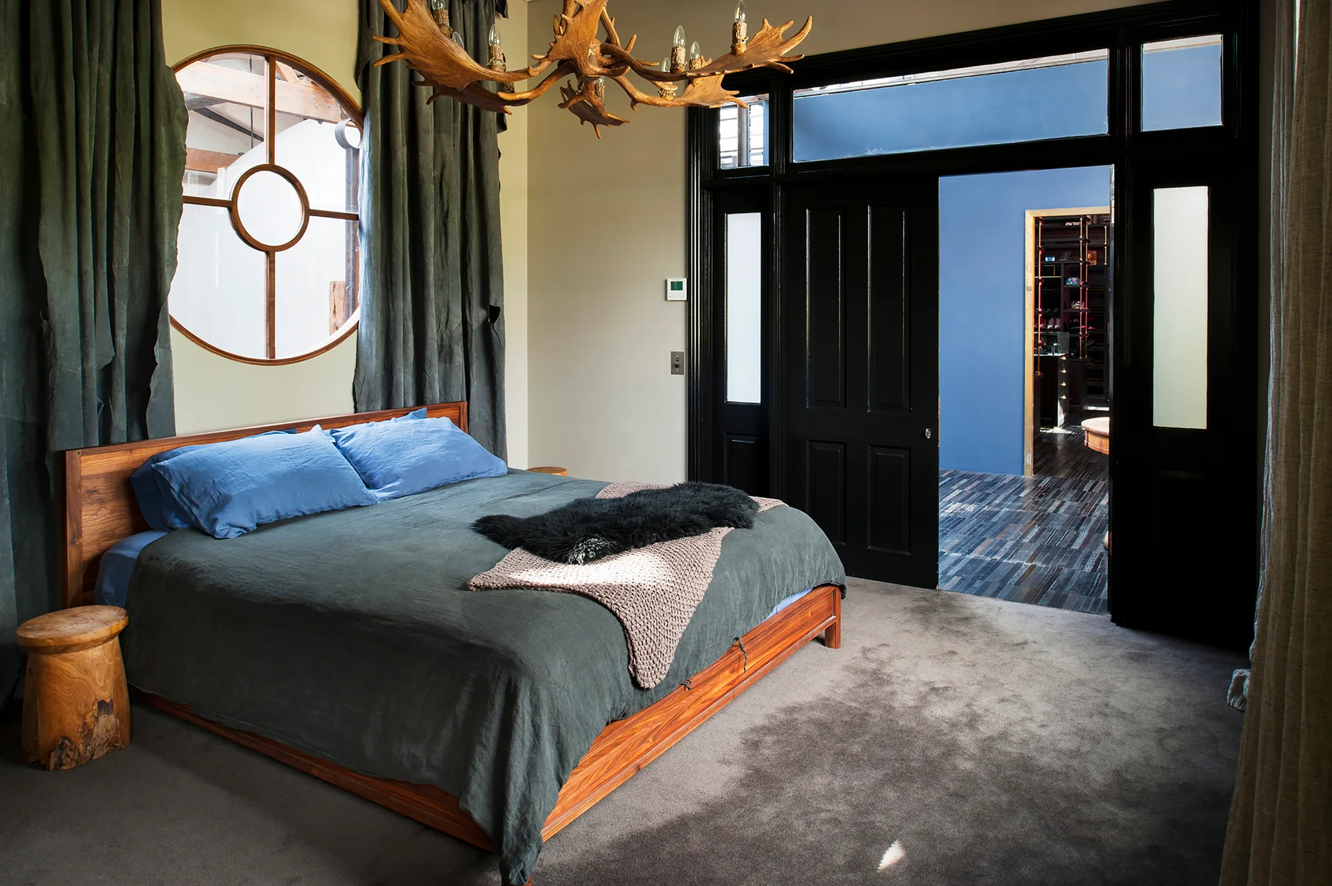 A rustic bedroom featuring a wooden bed, an antler chandelier overhead, and a circular window, with black doors opening to a blue walk-in closet.