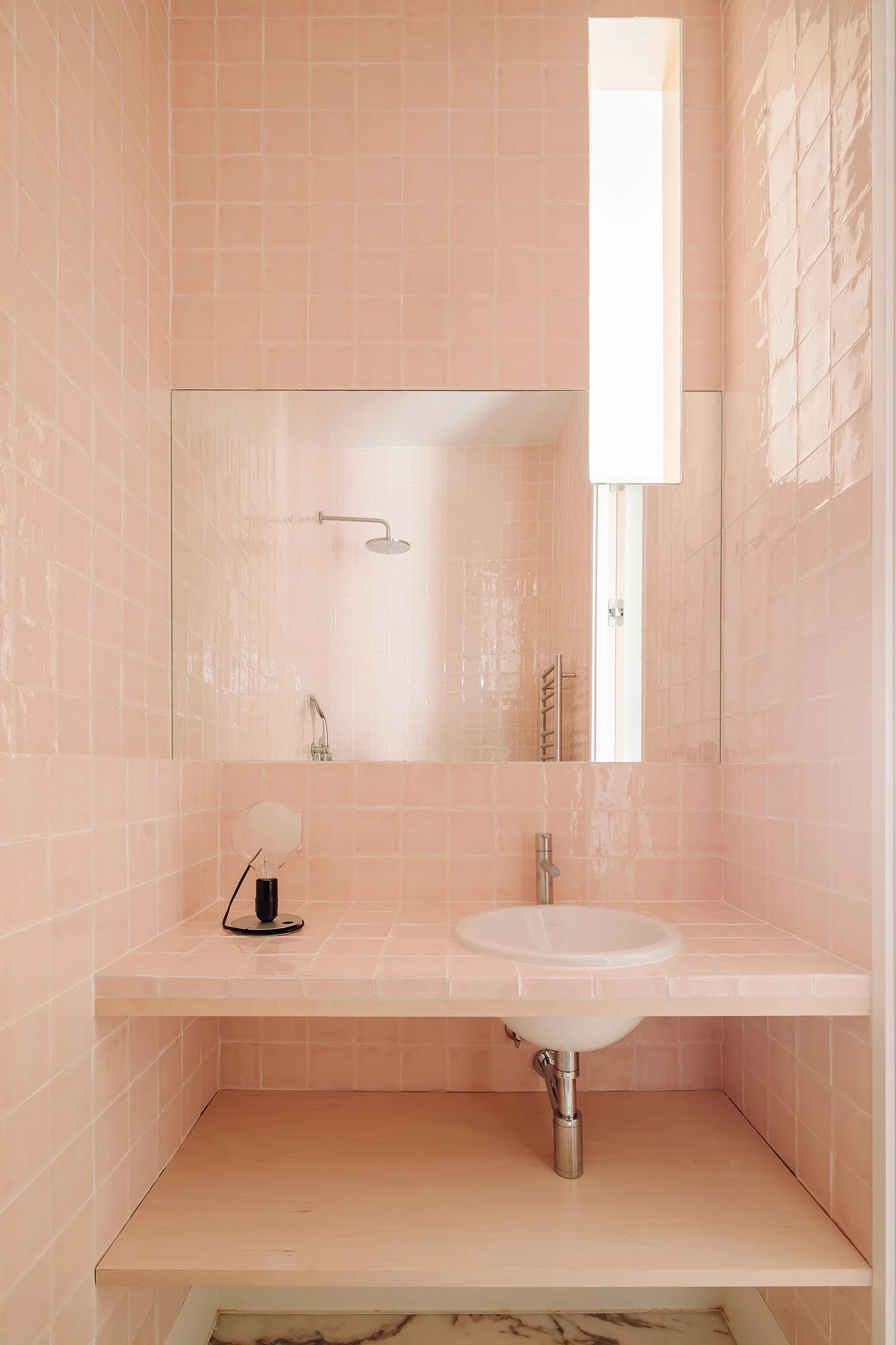 A minimalist bathroom with pink square tiled walls and vanity counter, a white basin sink, a wall mirror, and a light wood lower shelf.