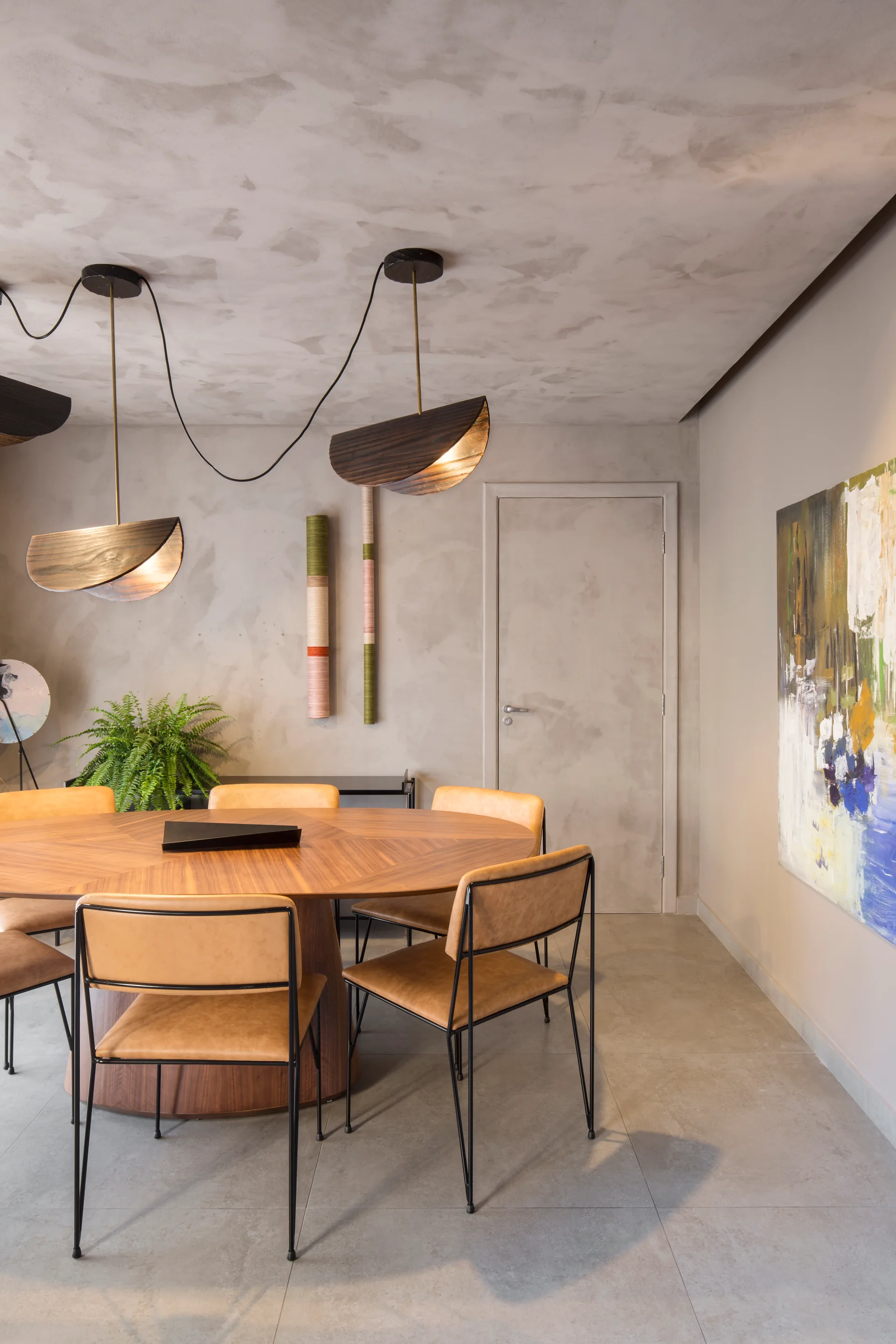 A contemporary dining room features a round wooden table, tan leather chairs with black frames, sculptural wood pendant lights, and textured grey walls.