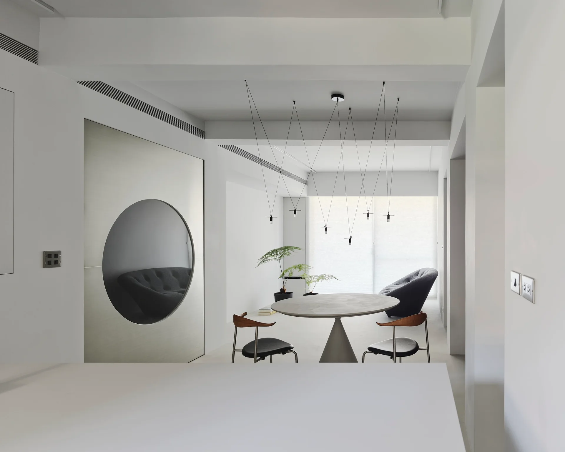 A minimalist dining room with a round pedestal table, black-seated chairs, and a silver metallic accent wall with a circular cutout.