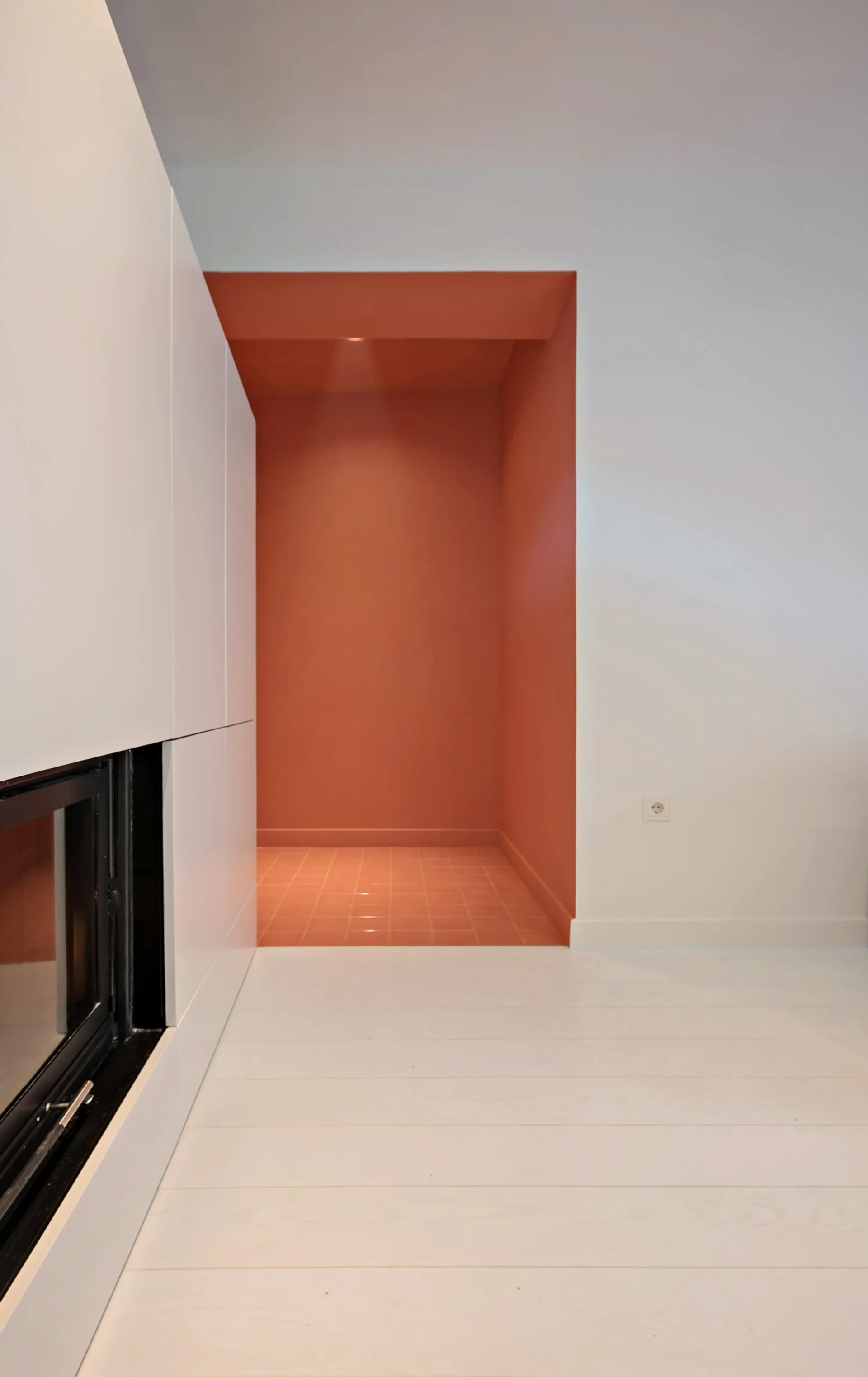 A minimalist white room features clean flat-front cabinetry next to a deeply recessed alcove clad in terracotta paint and floor tiles.
