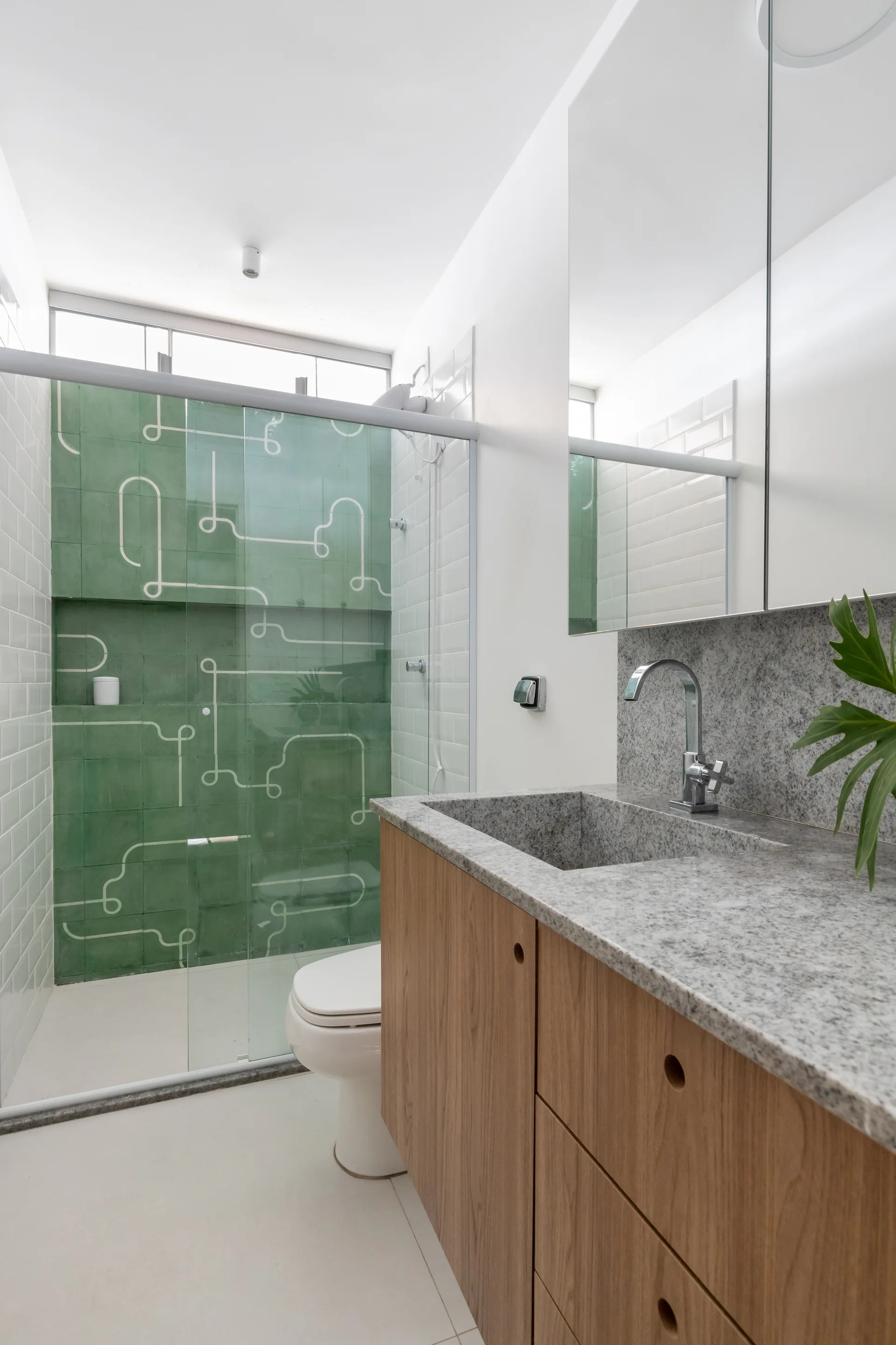 A modern bathroom features a wood vanity with a grey granite countertop and a glass-enclosed shower tiled in patterned green and white.