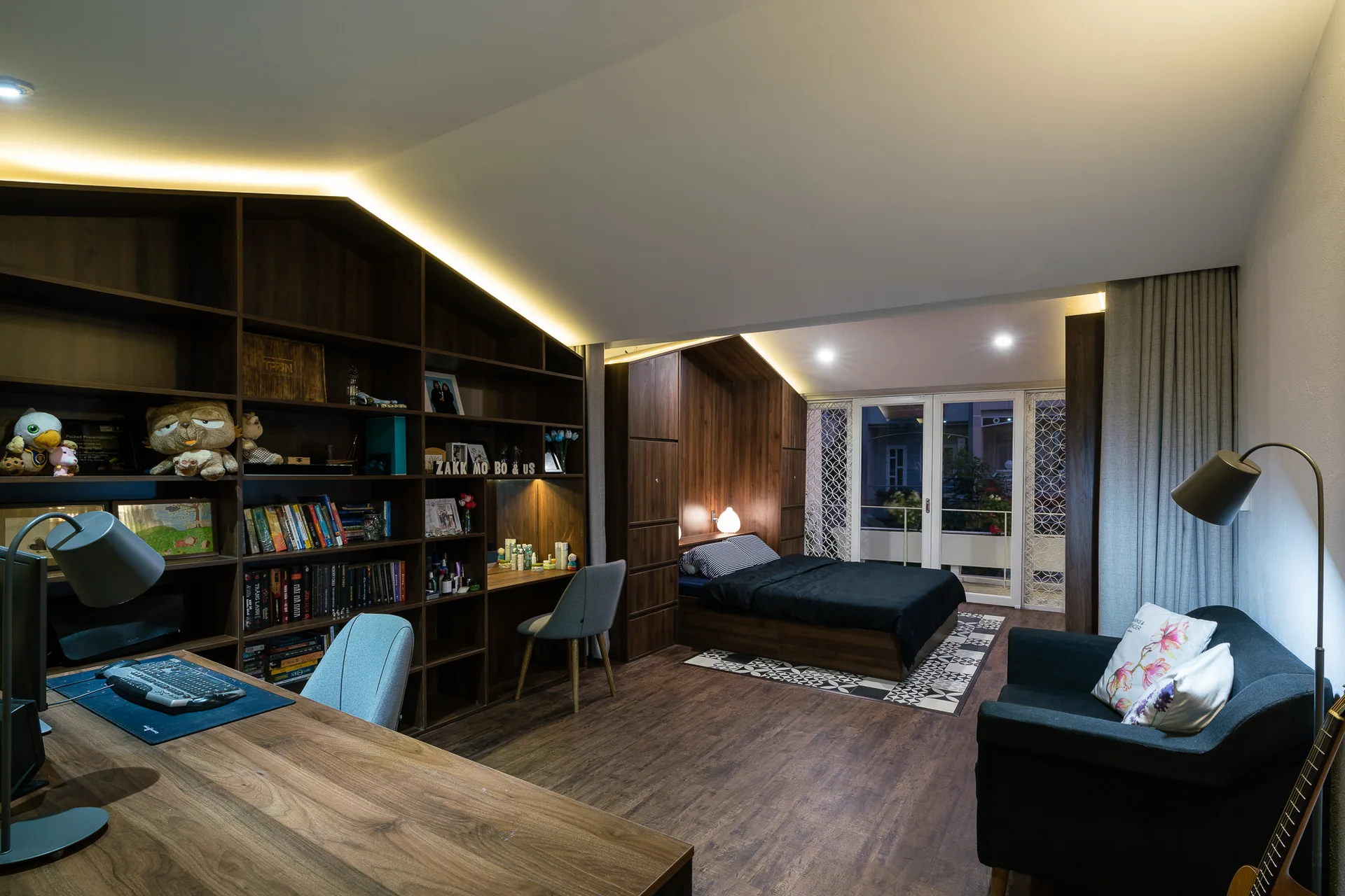 A modern multifunctional bedroom featuring a sloped ceiling, custom dark wood built-in shelving with desks, and a matching wood-paneled bed alcove.