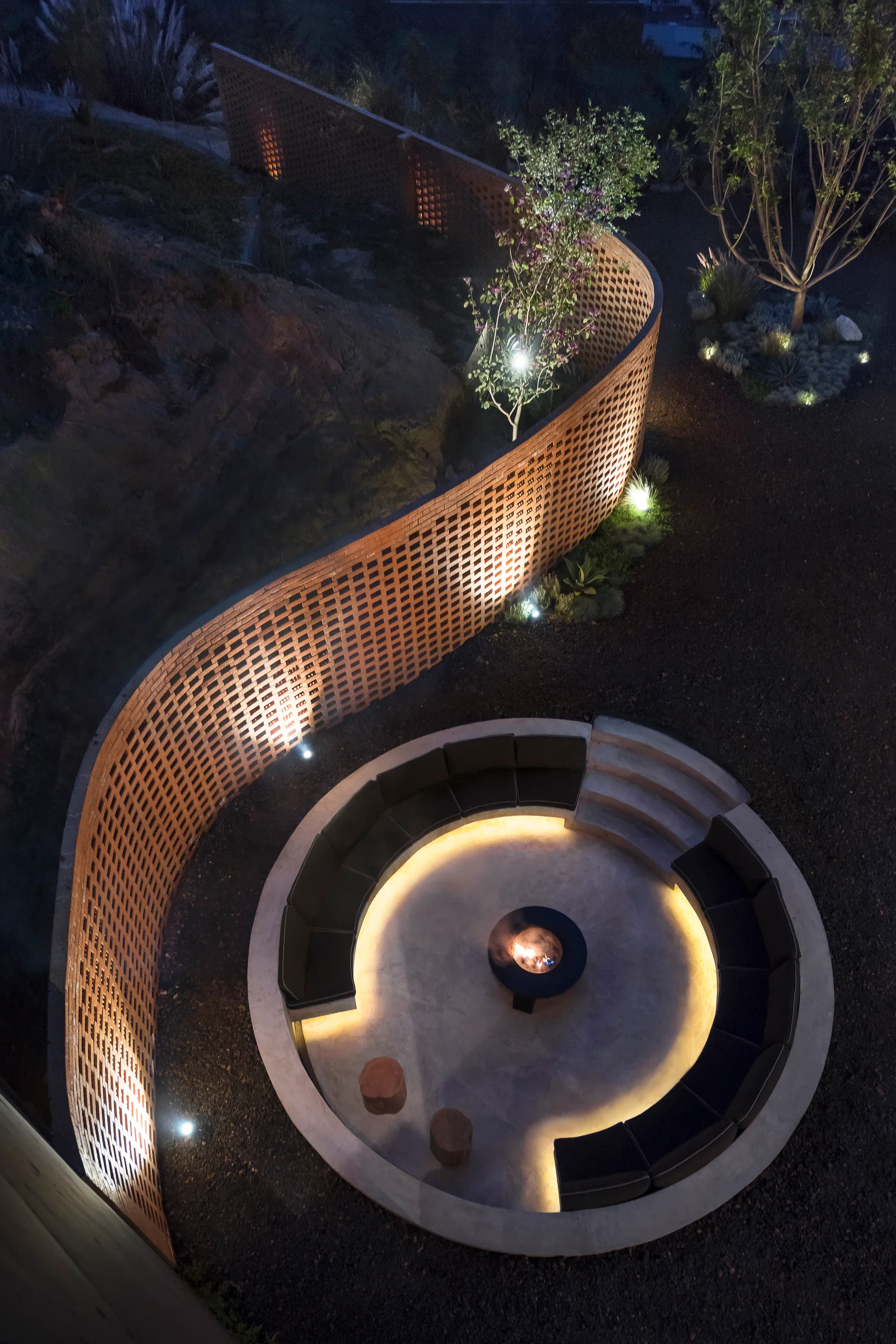 An aerial nighttime view of a circular sunken patio featuring built-in charcoal seating around a central fire pit, enclosed by a curved brick wall.