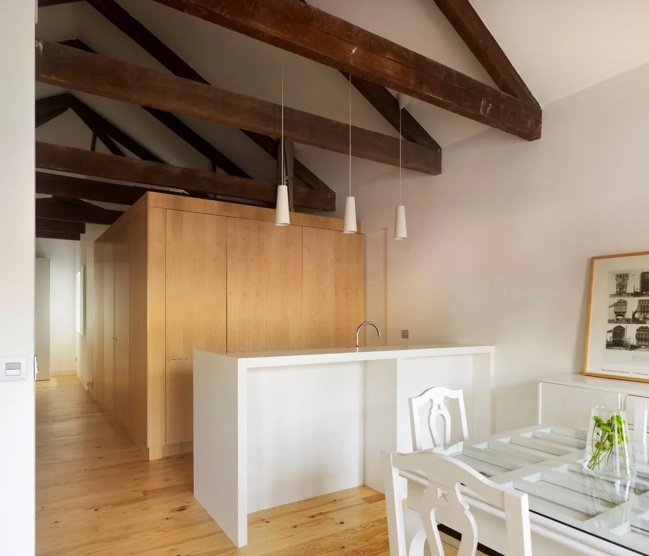 A bright open-plan kitchen features a minimalist white island beneath dark wood structural ceiling beams and a large timber storage cabinet.