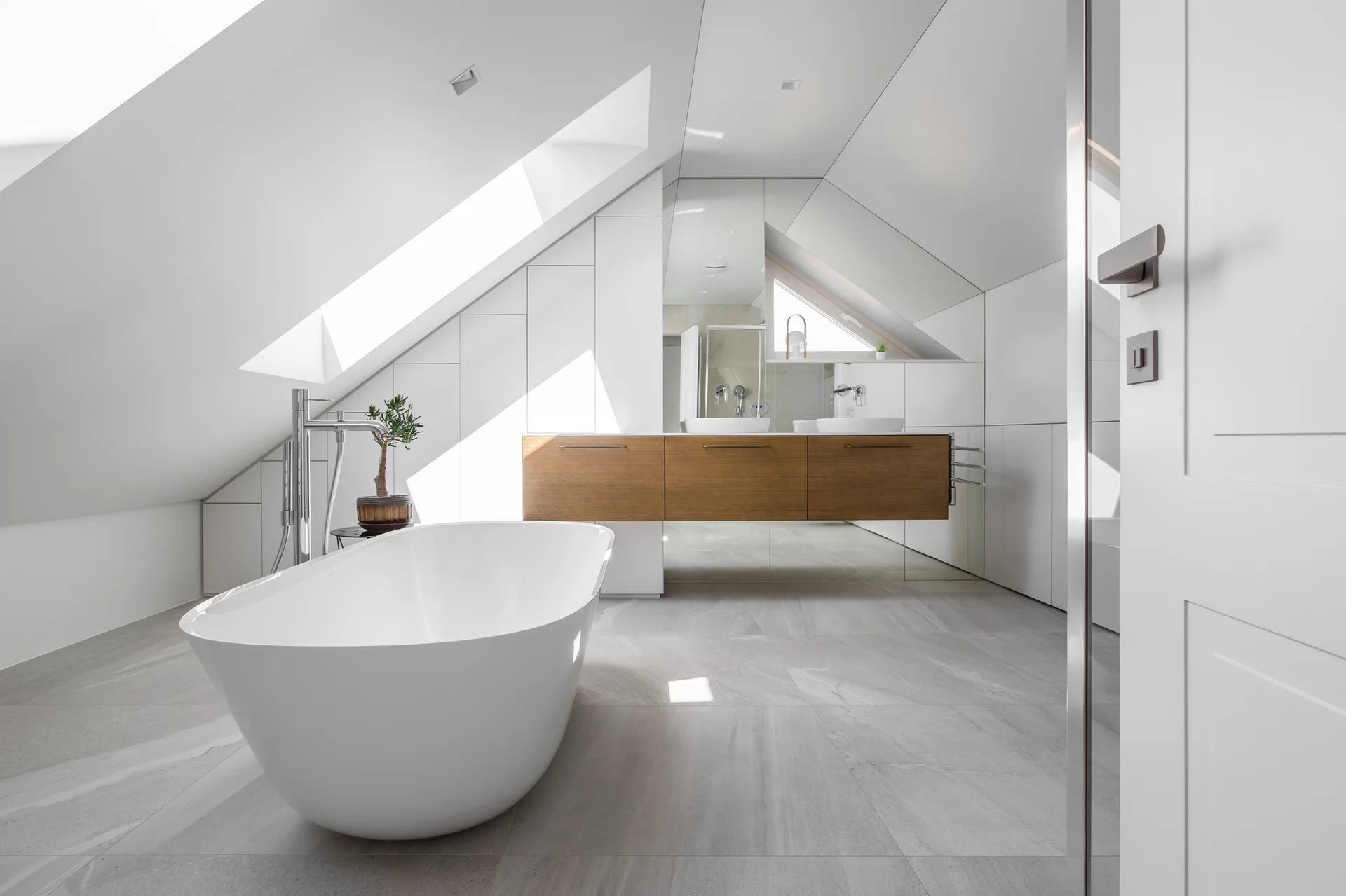 A modern attic bathroom featuring a white freestanding bathtub, a floating wood vanity with dual vessel sinks, and a sloped ceiling with a skylight.