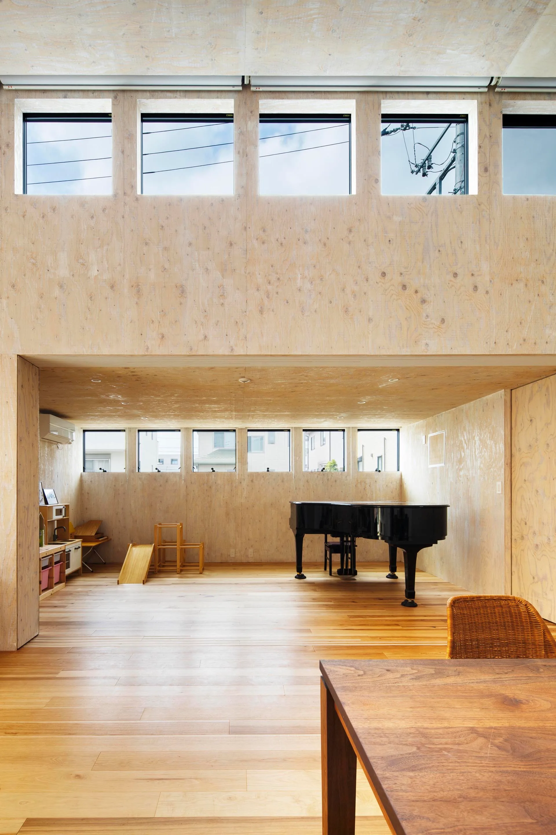 A spacious room finished in light plywood paneling features a black grand piano, small wooden children's toys, and double rows of square windows.