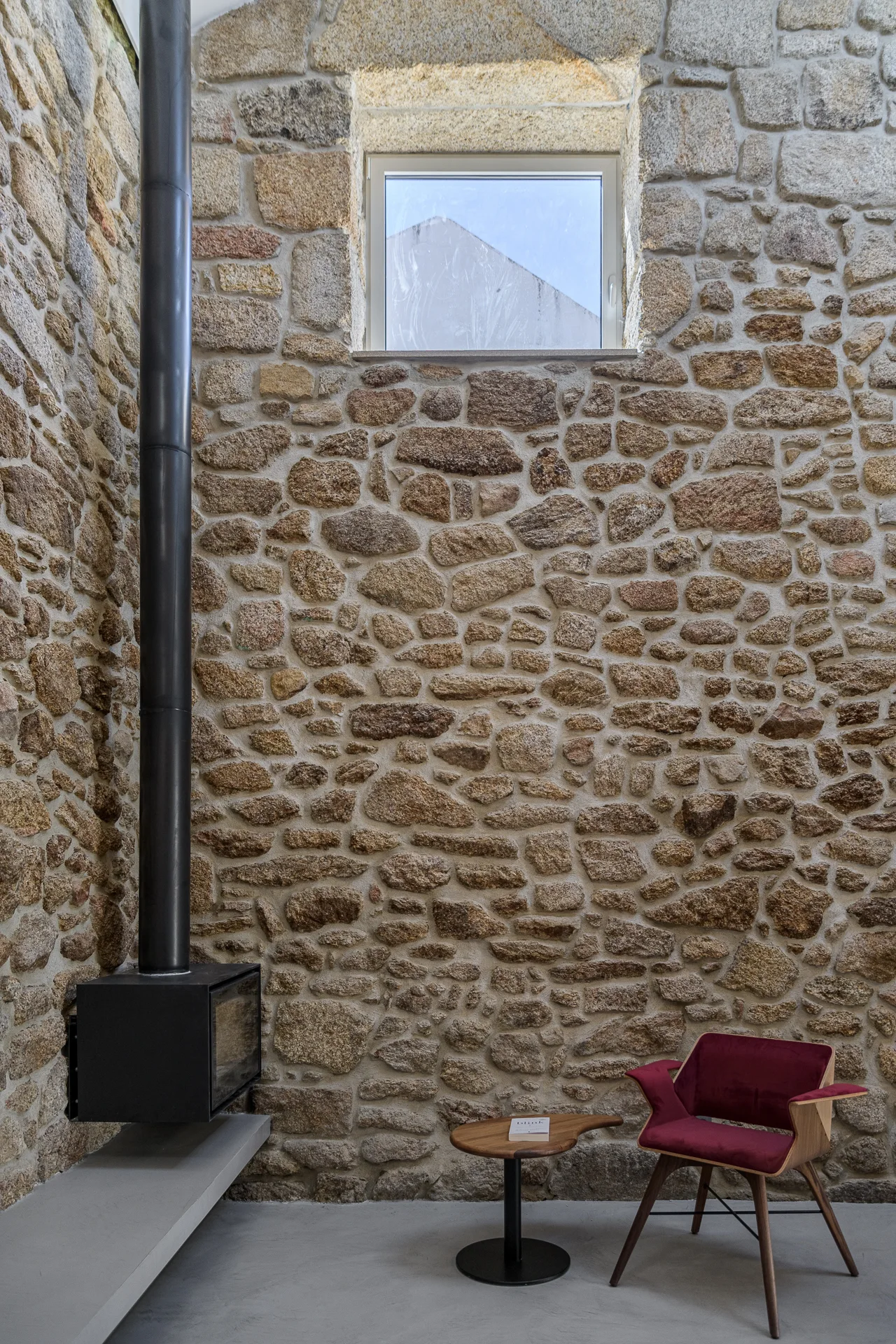A minimalist sitting area features an exposed stone masonry wall, a black metal fireplace, and a maroon armchair on a smooth concrete floor.