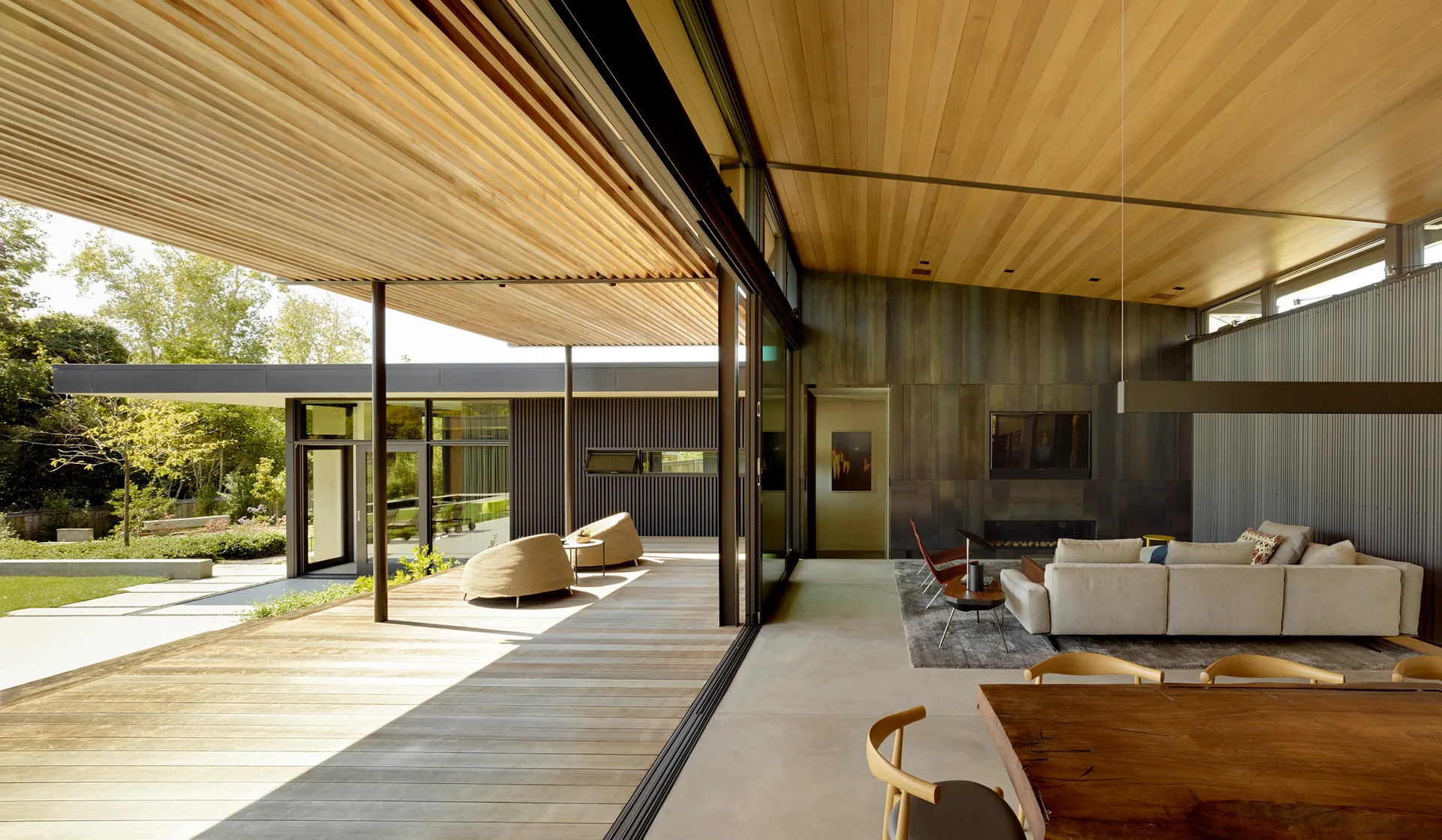 A modern living room with a cream sectional sofa and concrete floors opens onto a wooden deck through a large sliding glass door.
