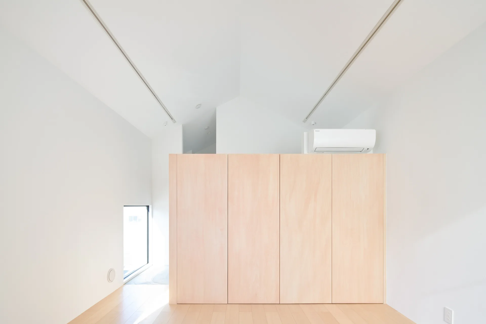A minimalist white room features a vaulted ceiling, light wood floors, a central plywood partition wall, and a small low window.