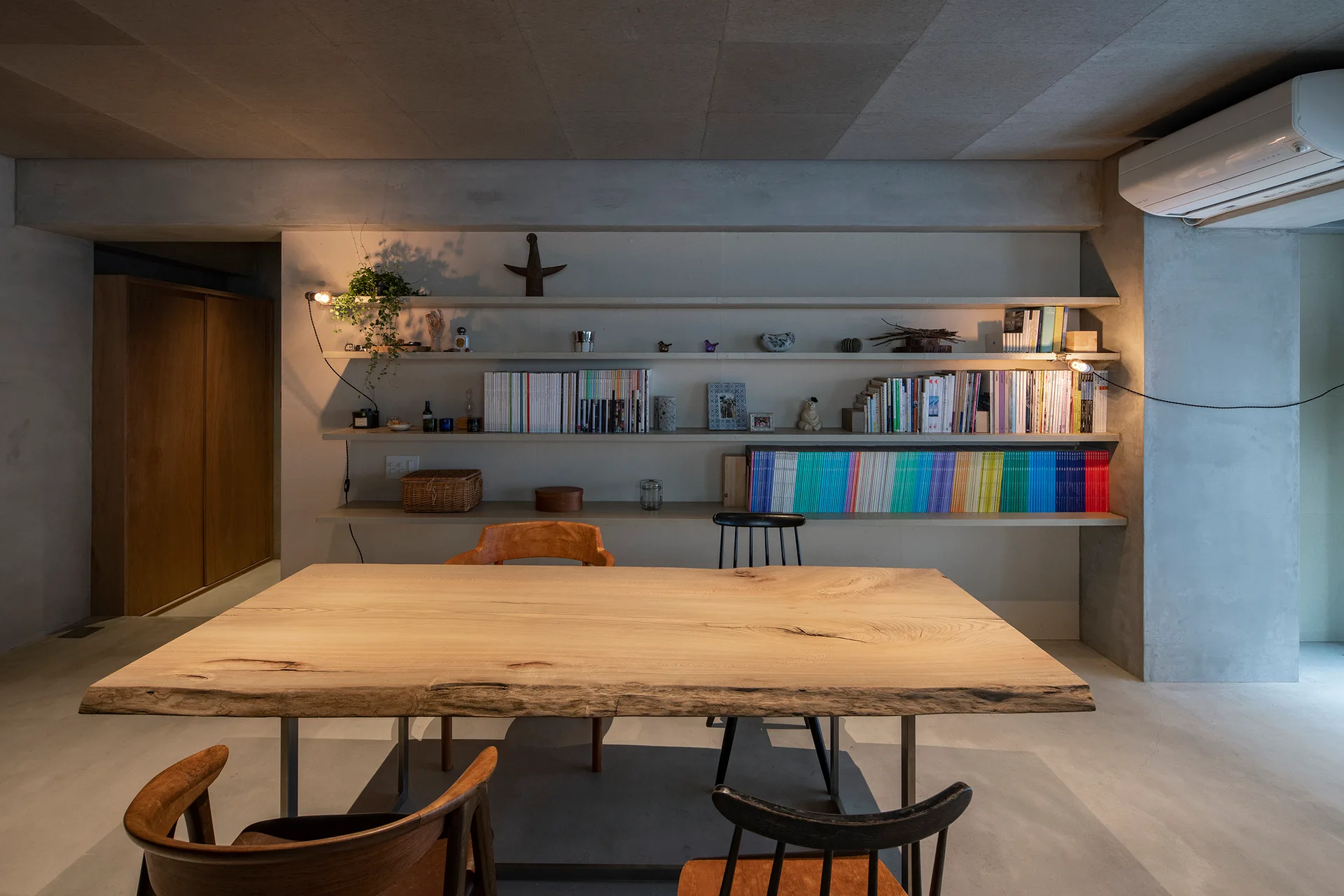 A minimalist dining room features a raw concrete wall and ceiling, a live-edge wooden table with chairs, and wall-mounted shelving filled with books.