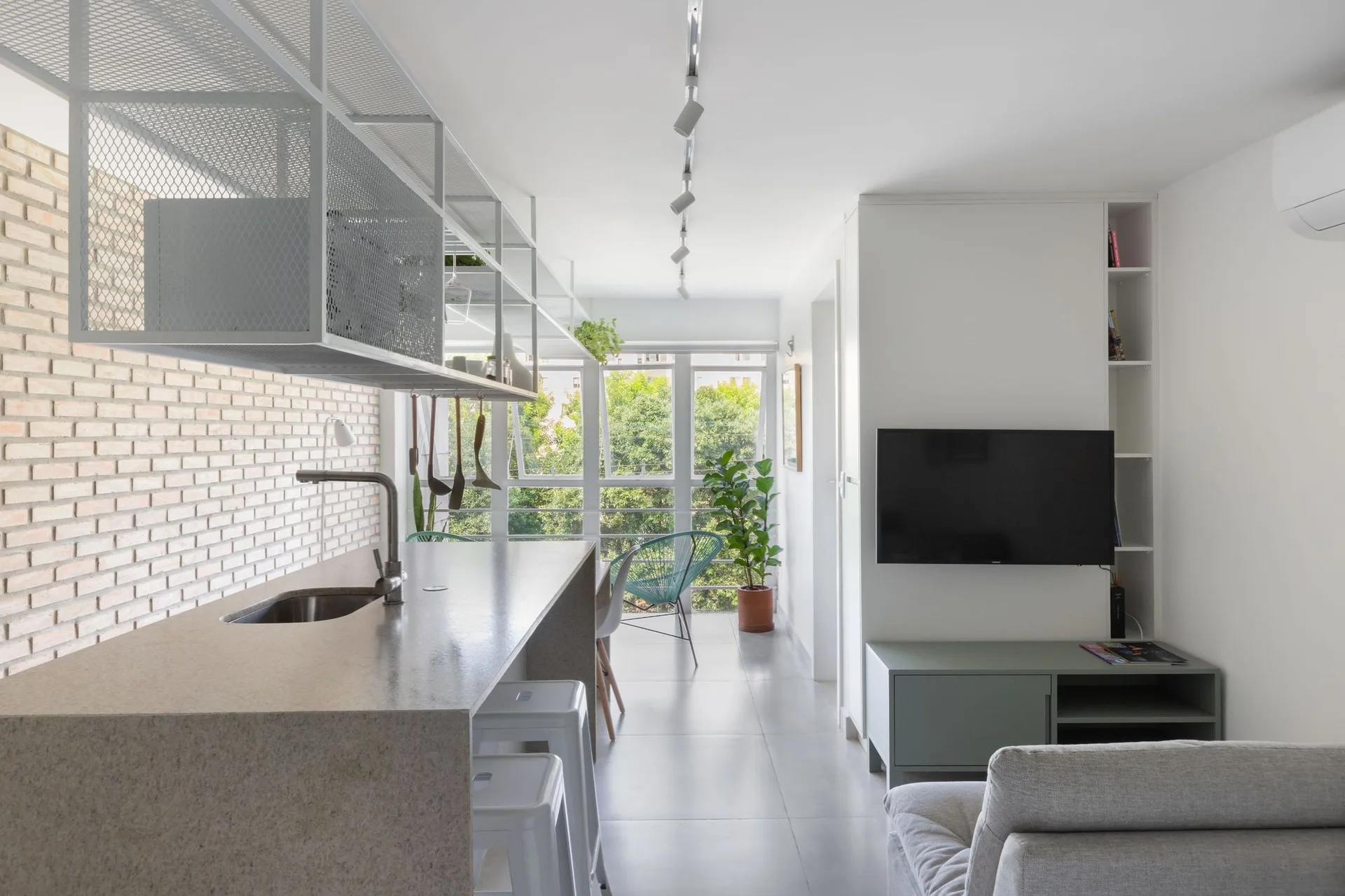 An open-concept kitchen and living area features a stone island with white stools, a white painted brick wall, and a hanging white mesh shelf.