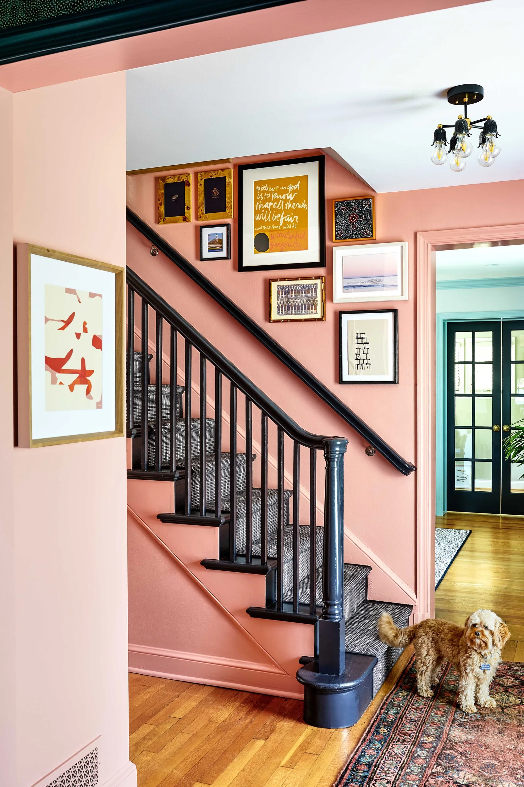 A wooden staircase with a black railing and dark patterned runner stands against pink walls decorated with a diagonal gallery wall of framed art.
