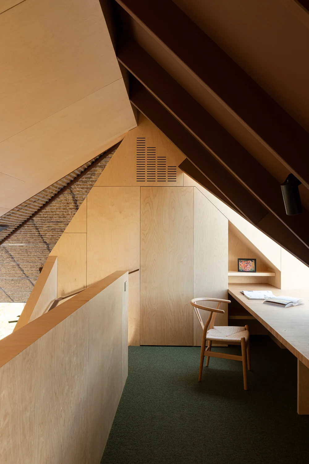 A minimalist attic office features light plywood paneling, a built-in wooden desk with a woven chair, and dark green carpeting under a sloped ceiling.