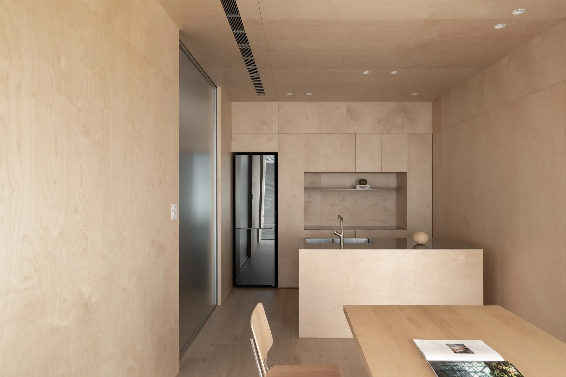 A minimalist kitchen and dining area features seamless light plywood paneling on the walls and ceiling, a central island, and a wooden dining table.