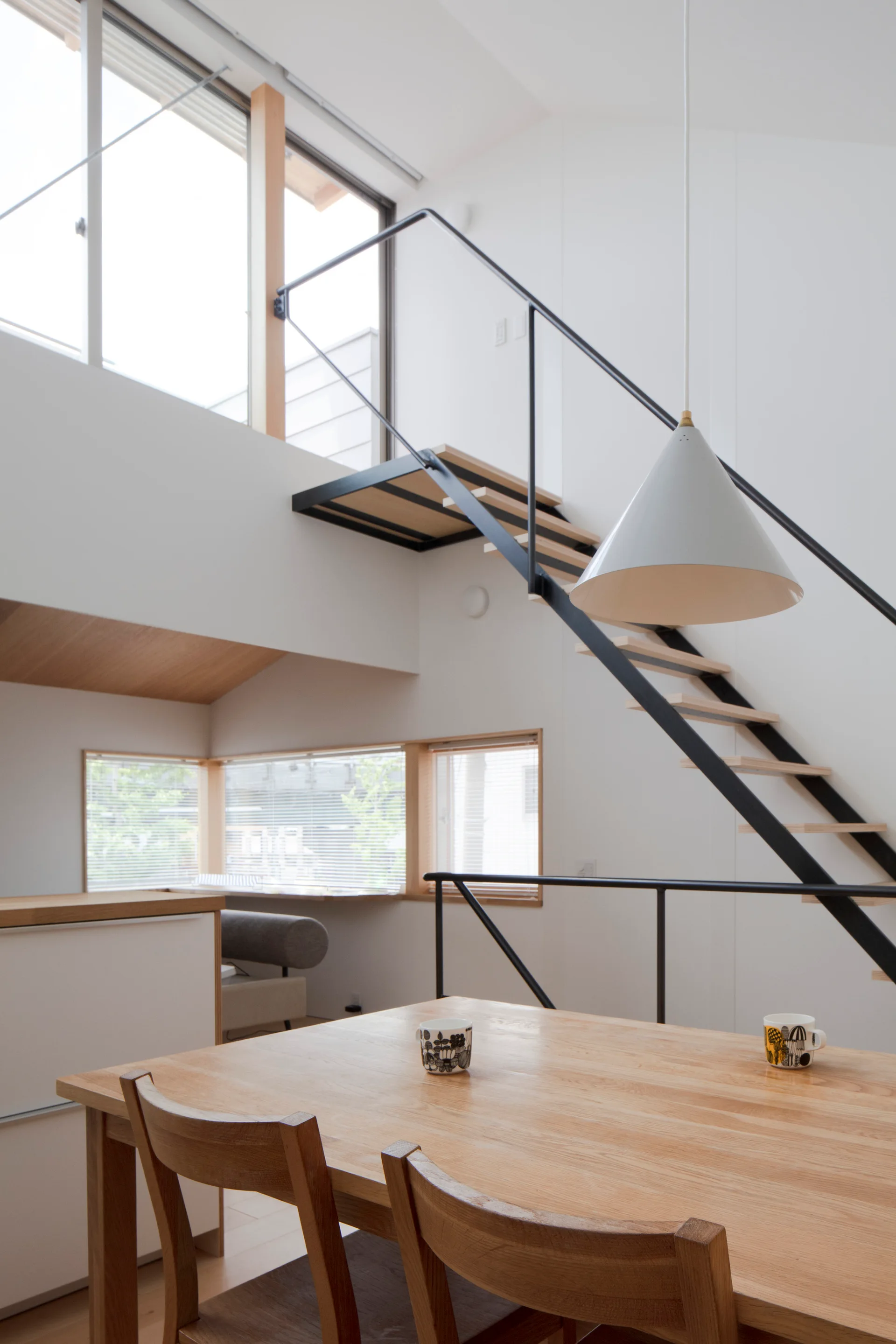 A minimalist dining room features a light wood table, wooden chairs, a white pendant light, and a floating staircase with a black metal handrail.