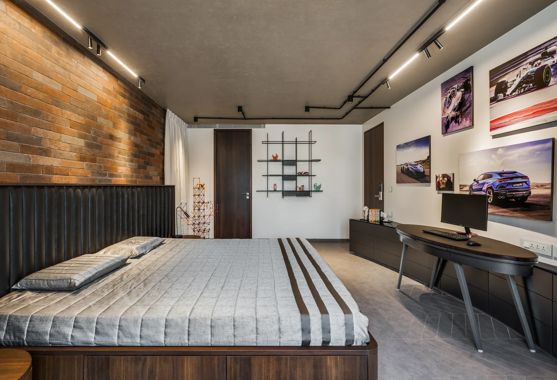 A modern bedroom featuring an exposed brick accent wall behind a platform bed, a curved dark wood desk, and black track lighting on a grey ceiling.
