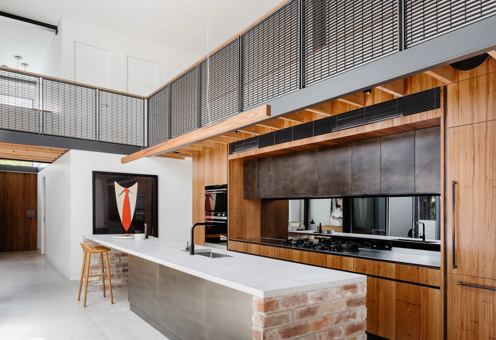 A modern industrial kitchen featuring a brick-base island, wood cabinets, a polished concrete floor, and an overhead mezzanine loft with mesh railing.