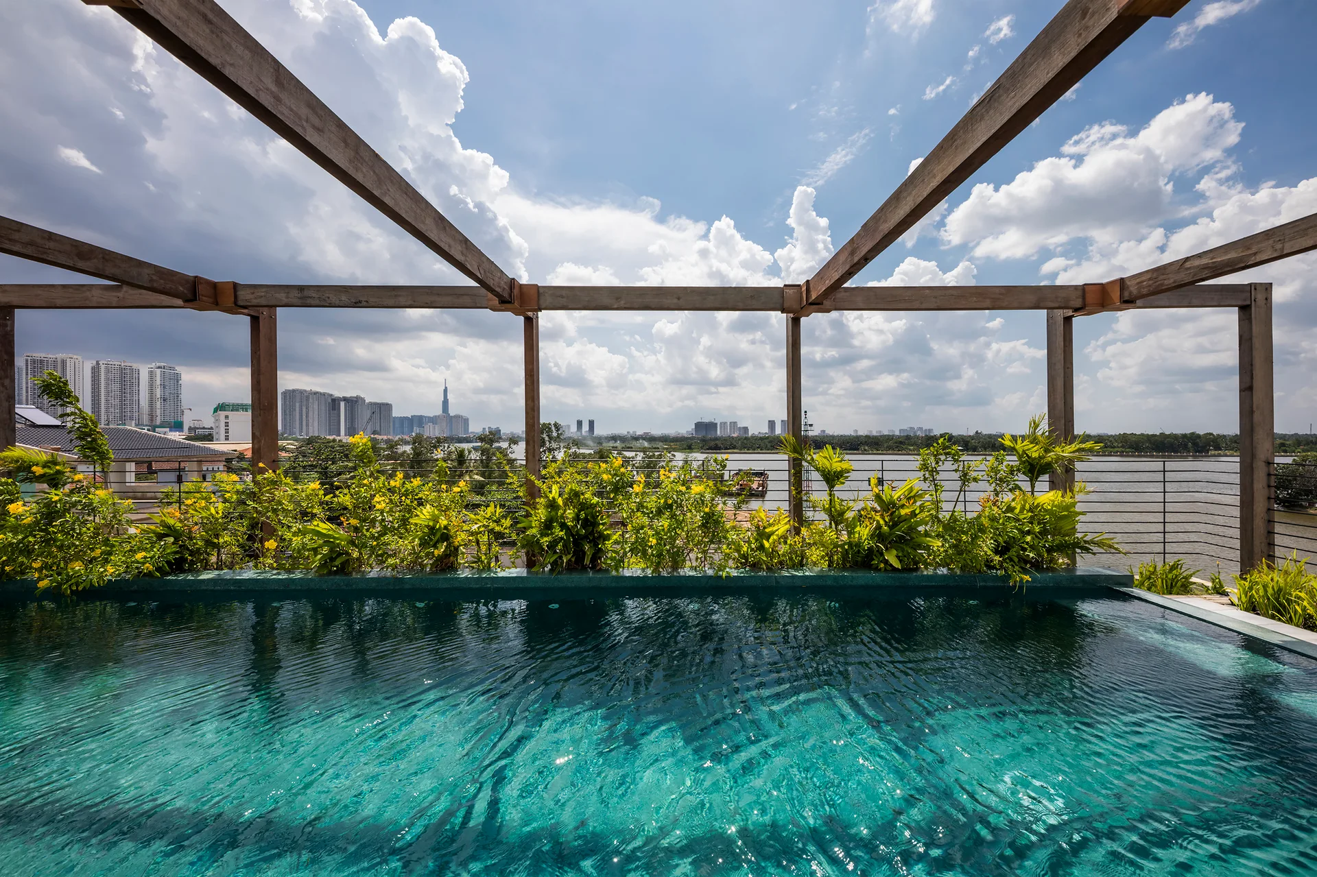 A turquoise infinity pool on a rooftop terrace features a weathered wood pergola and green plants overlooking a distant city and river.