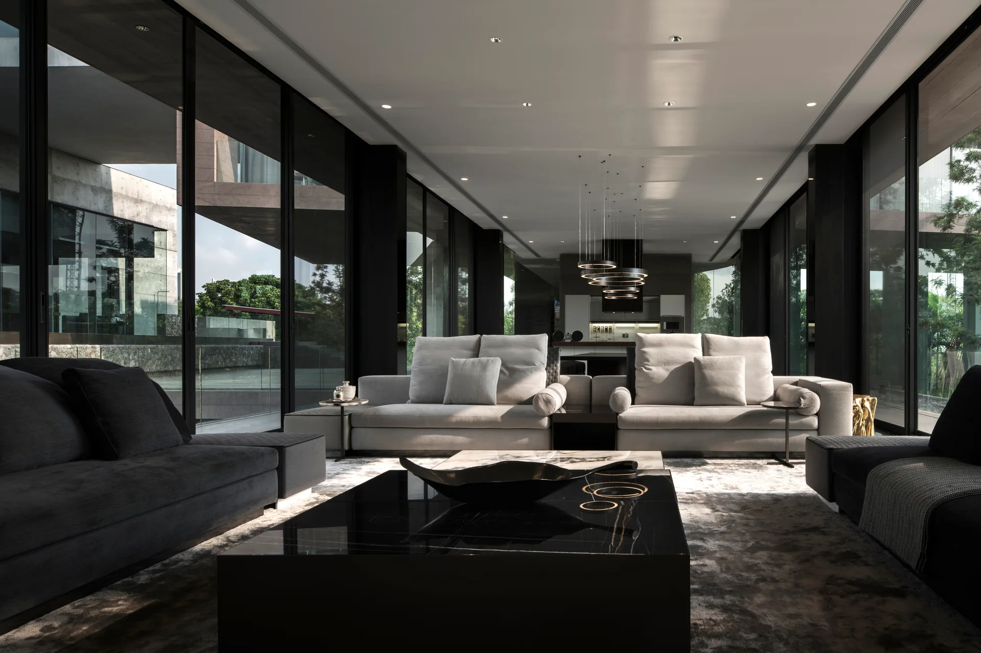 A modern minimalist living room featuring symmetrical light grey sofas and a large glossy black coffee table surrounded by floor-to-ceiling glass walls.