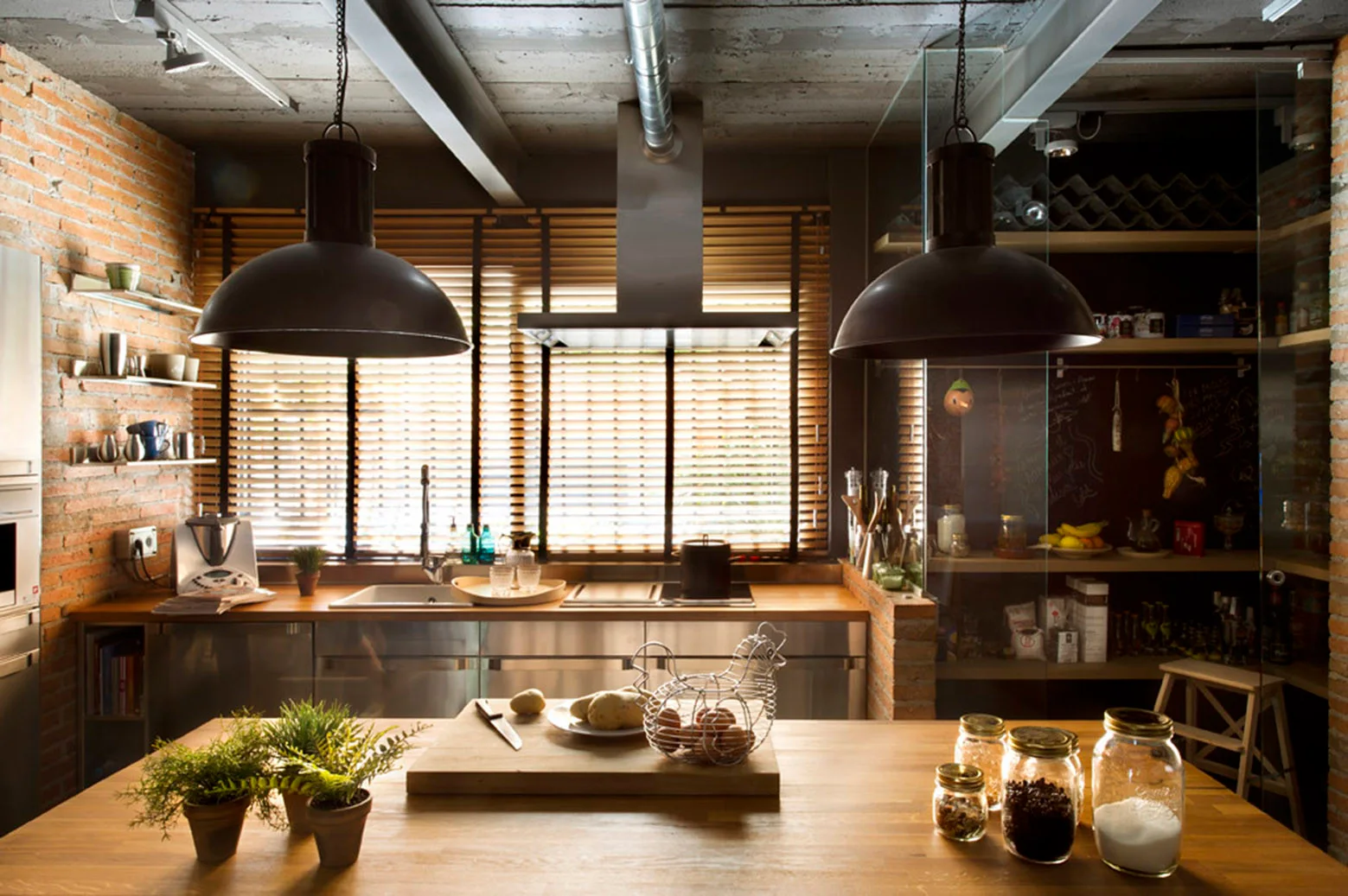 An industrial kitchen featuring a wooden island with potted plants, stainless steel countertops under wooden blinds, and a glass-enclosed pantry.