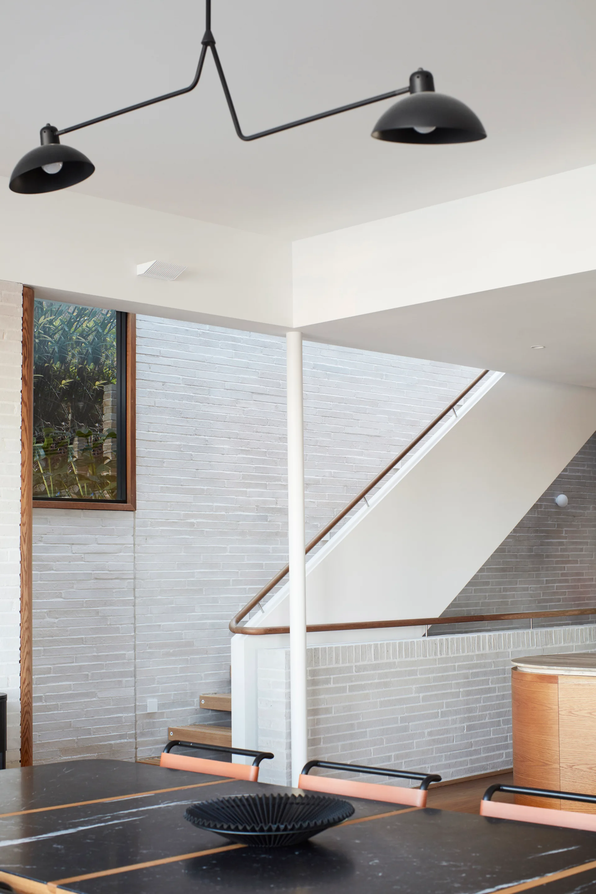 A modern staircase features a white brick wall and wooden handrail, with a black dining table and minimalist pendant light in the foreground.