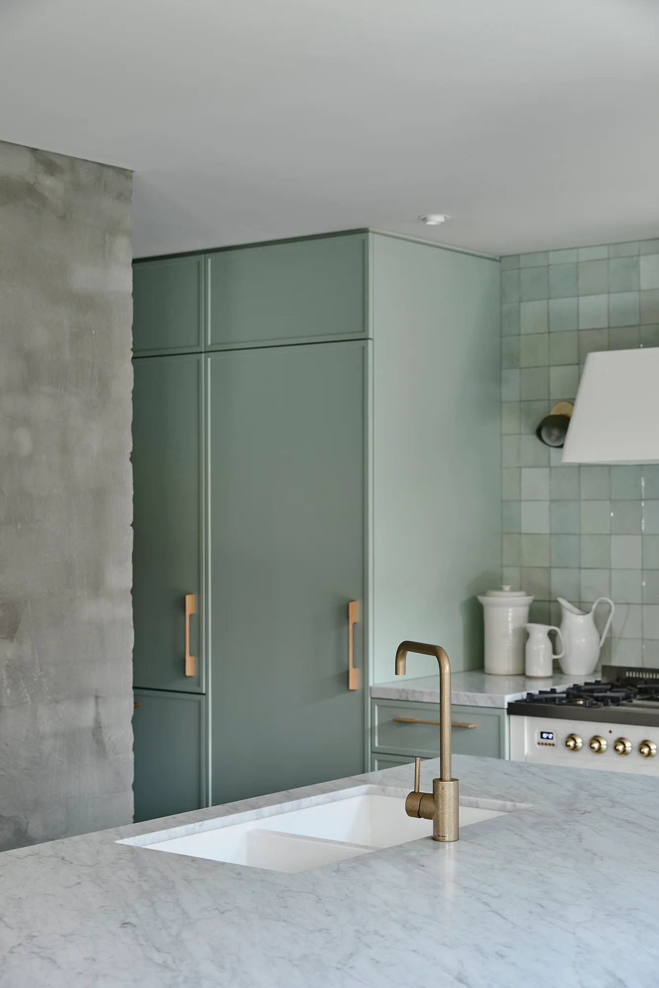 A modern kitchen featuring sage green cabinetry, a matching square tile backsplash, and a marble island with a brass faucet.