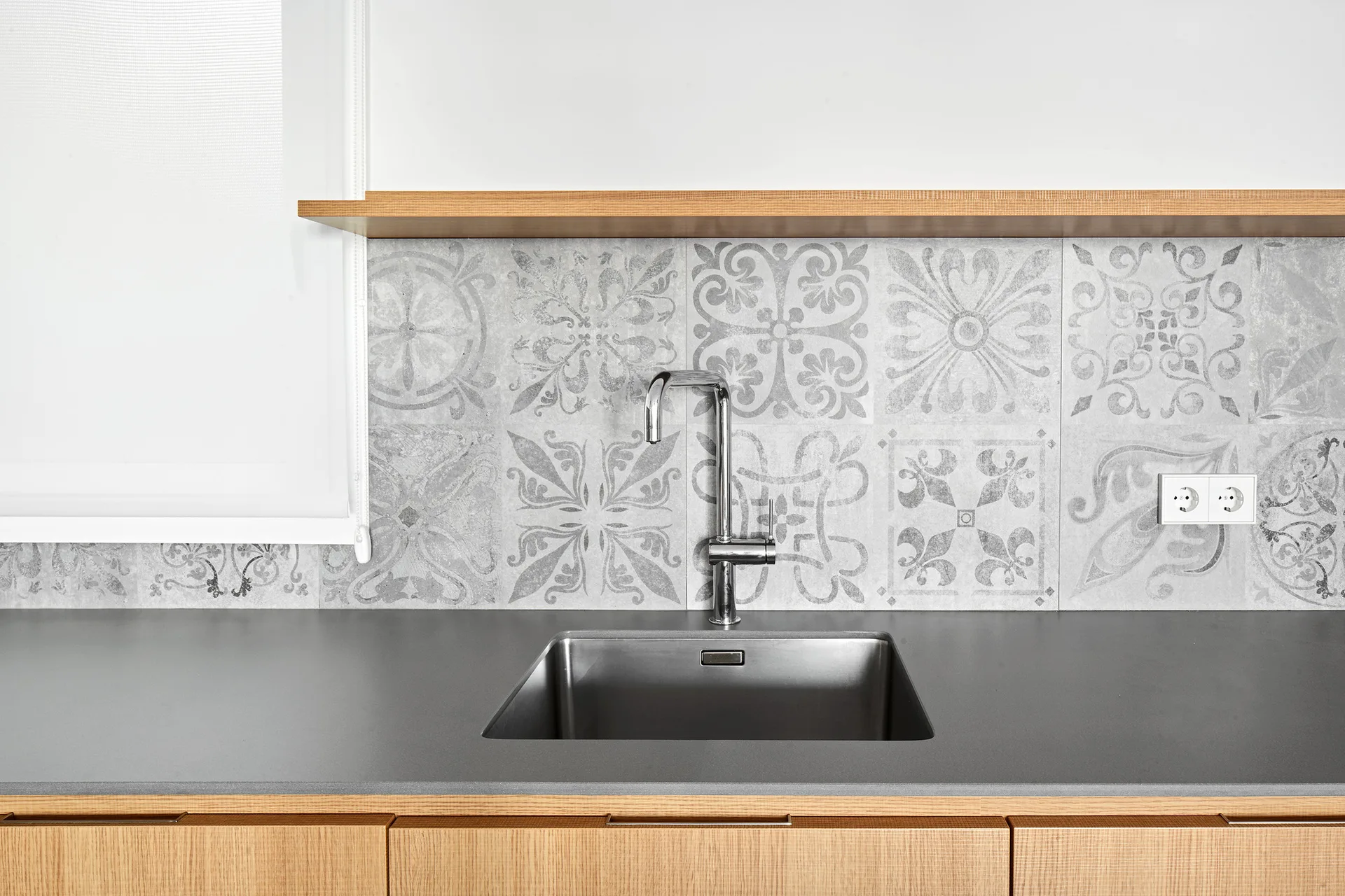A modern kitchen sink with a chrome faucet is built into a gray countertop beneath a patterned gray tile backsplash and a wood floating shelf.