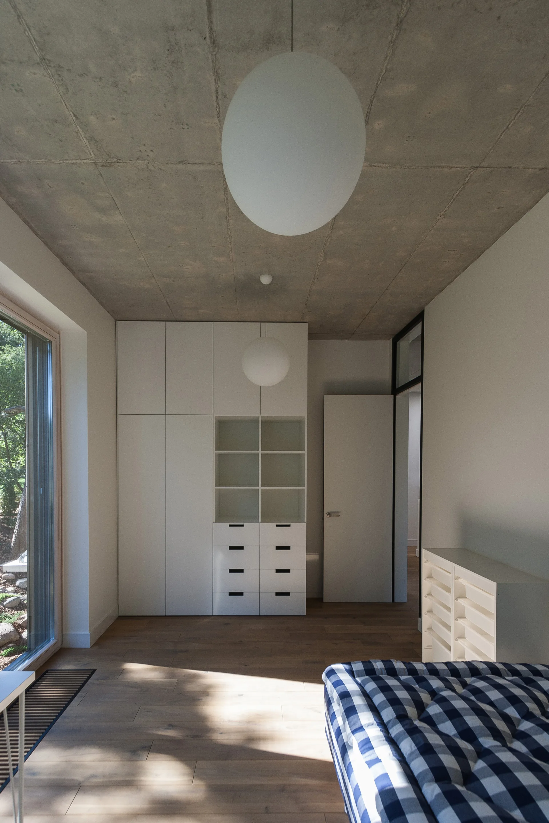 A minimalist bedroom featuring a raw concrete ceiling, white built-in wardrobe, natural wood flooring, and a blue and white checkered bed.