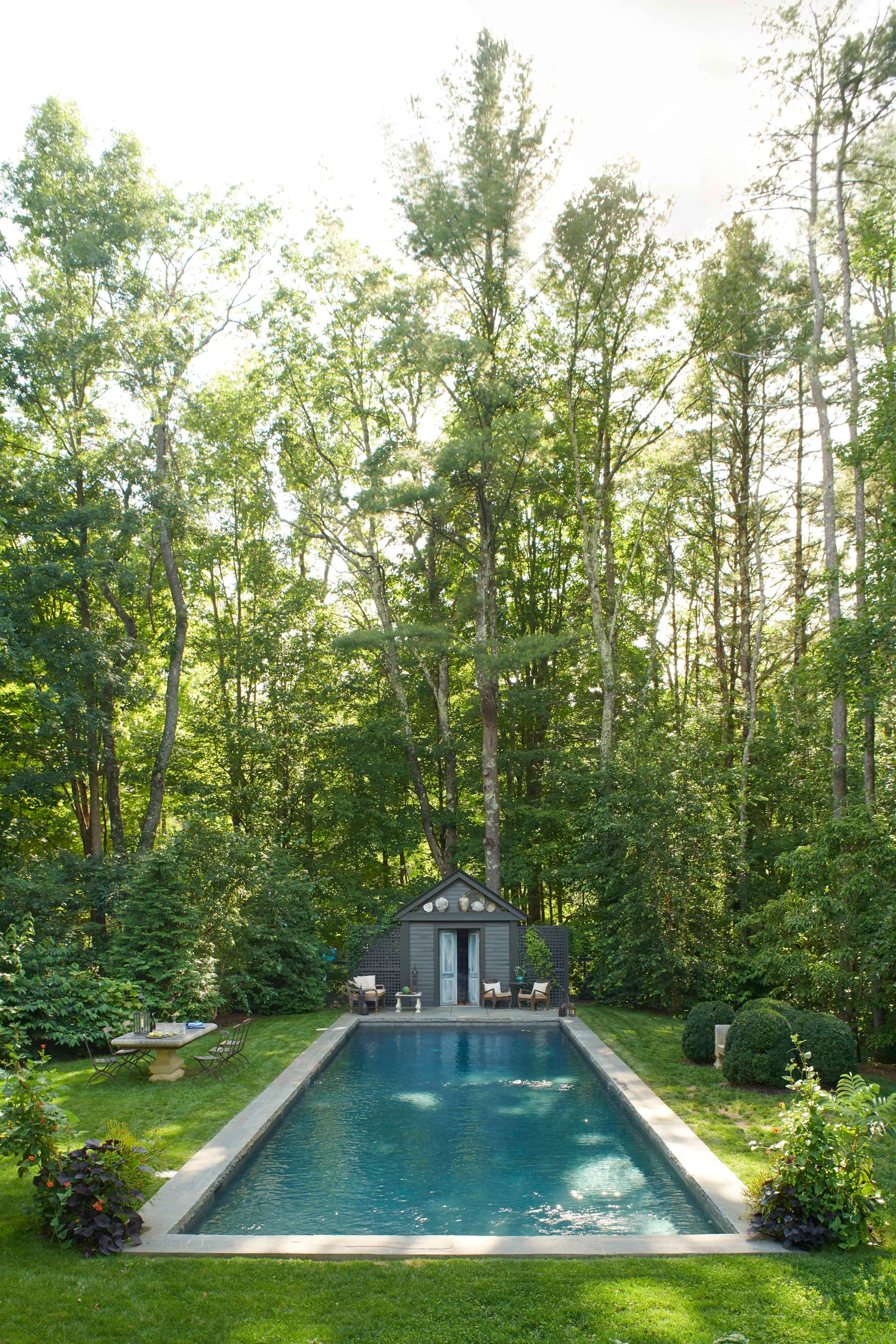A rectangular swimming pool surrounded by a green lawn leads to a small gray pool house nestled in a dense forest.