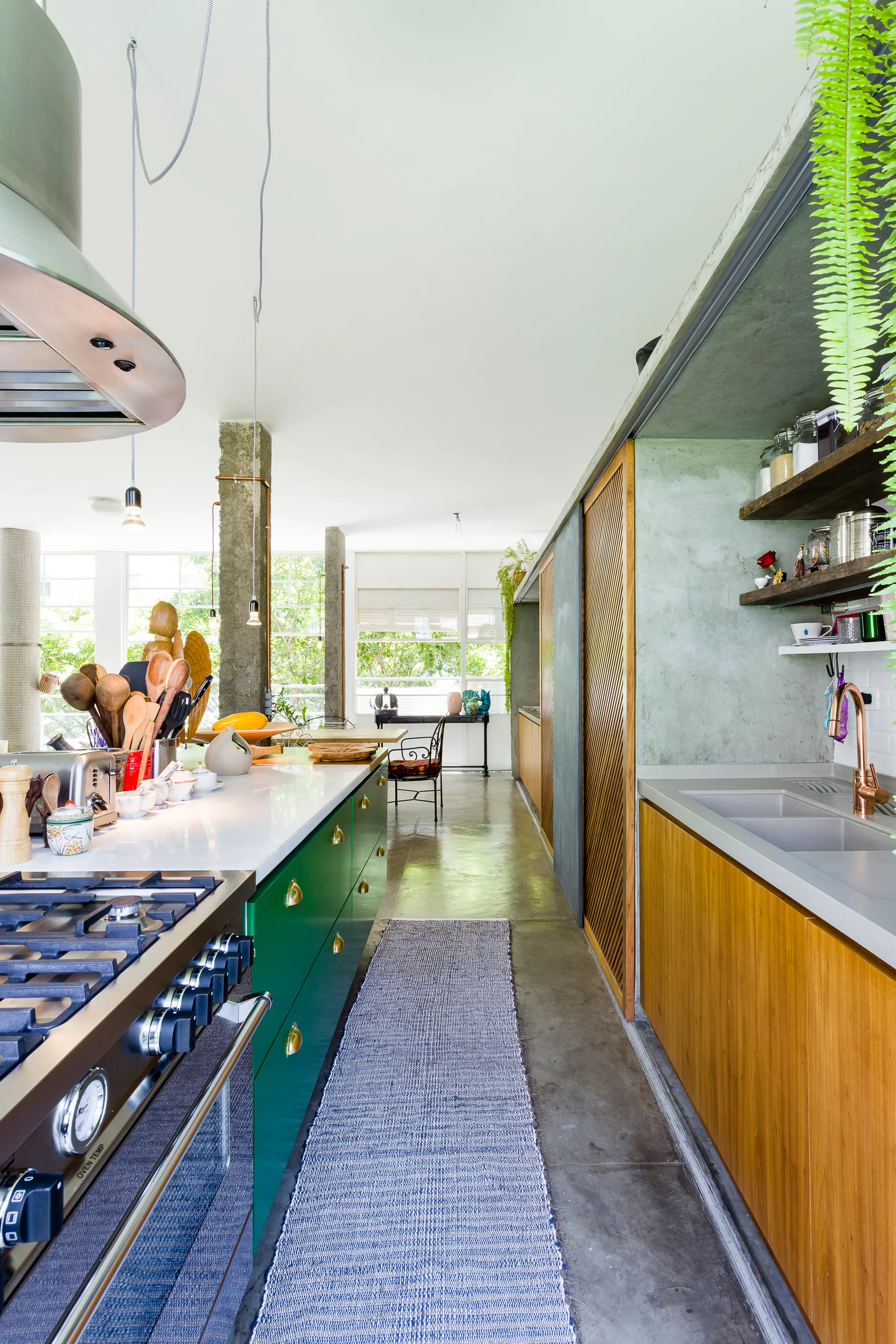 A galley kitchen features a green painted island with a metal range, warm wood cabinetry, polished concrete floors, and a blue runner rug.