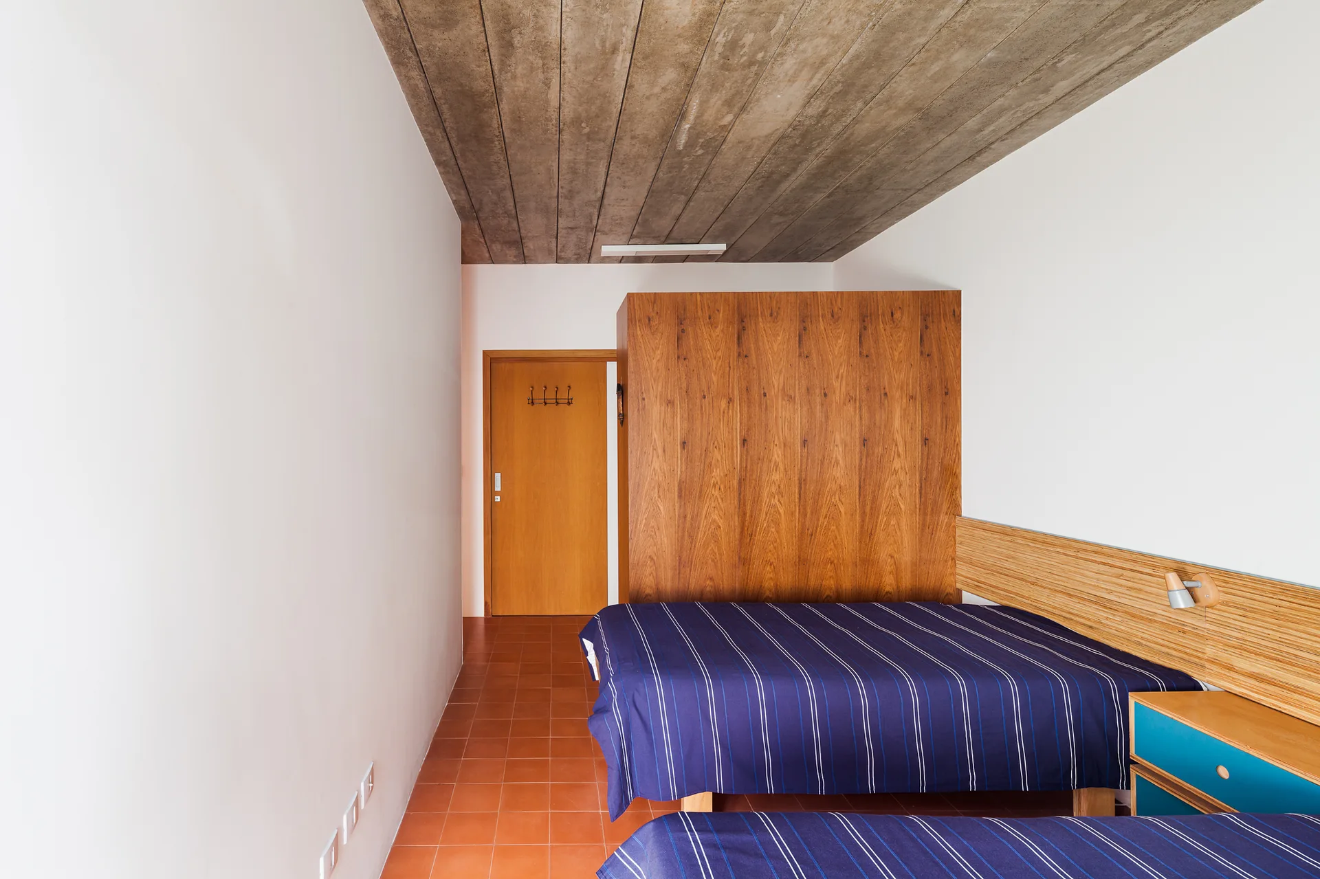 A minimalist bedroom featuring two beds with blue striped covers, a wood storage partition, terracotta floor tiles, and an exposed concrete ceiling.