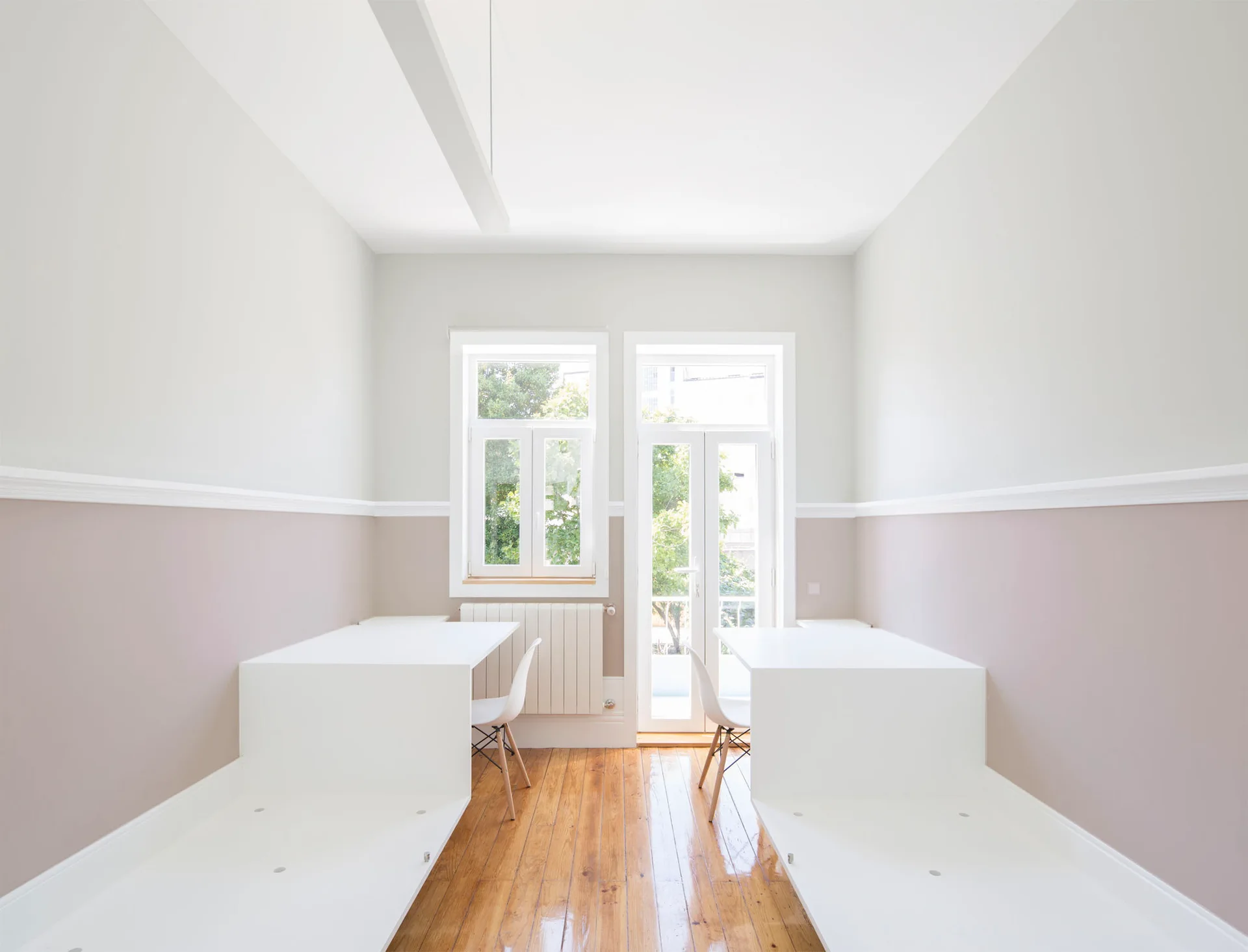 A symmetrical minimalist home office with two custom white desks, warm hardwood floors, and walls split into mauve and white sections.