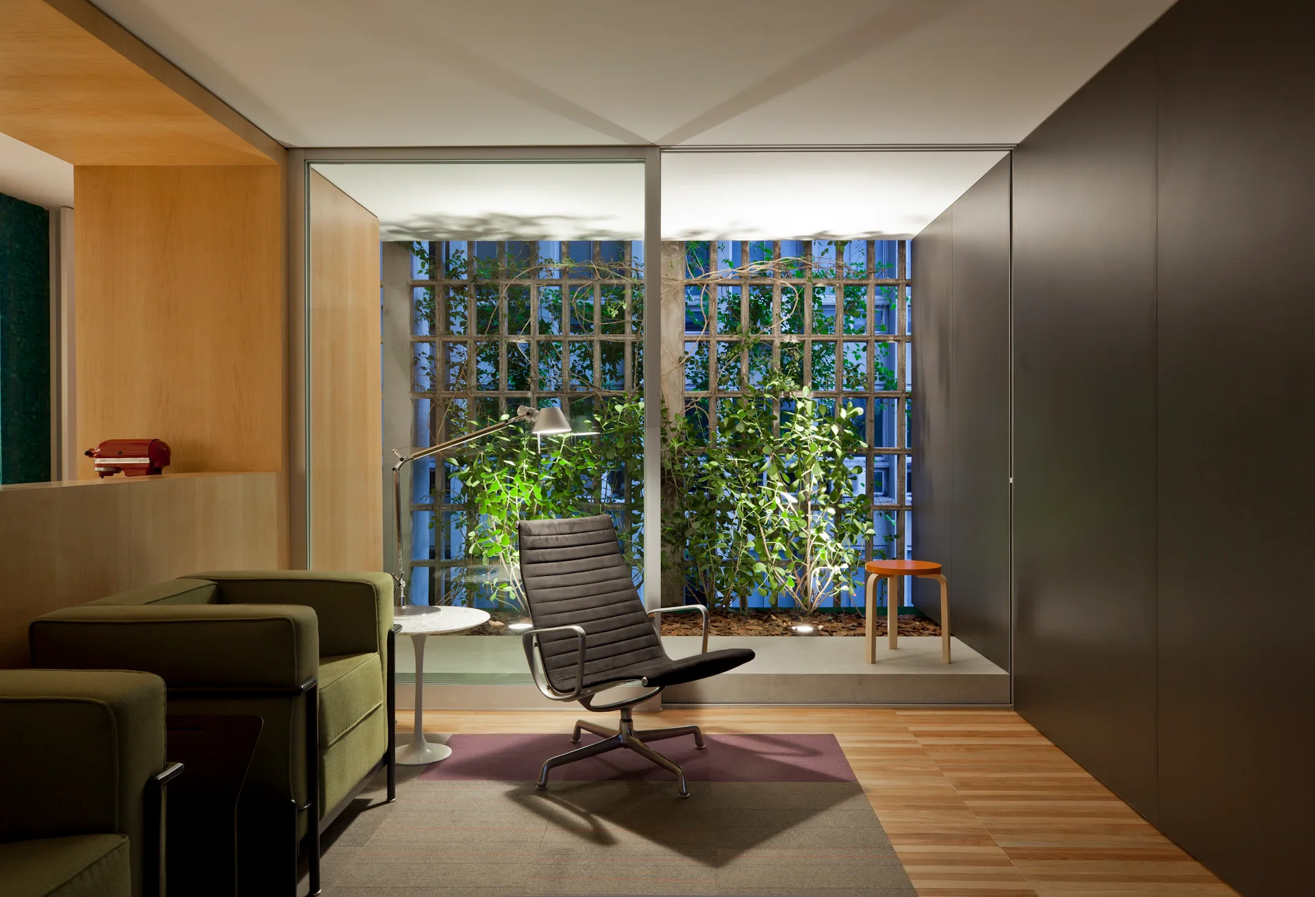 A modern lounge room features a green armchair and a black ribbed leather chair in front of a floor-to-ceiling glass wall showcasing a green courtyard.
