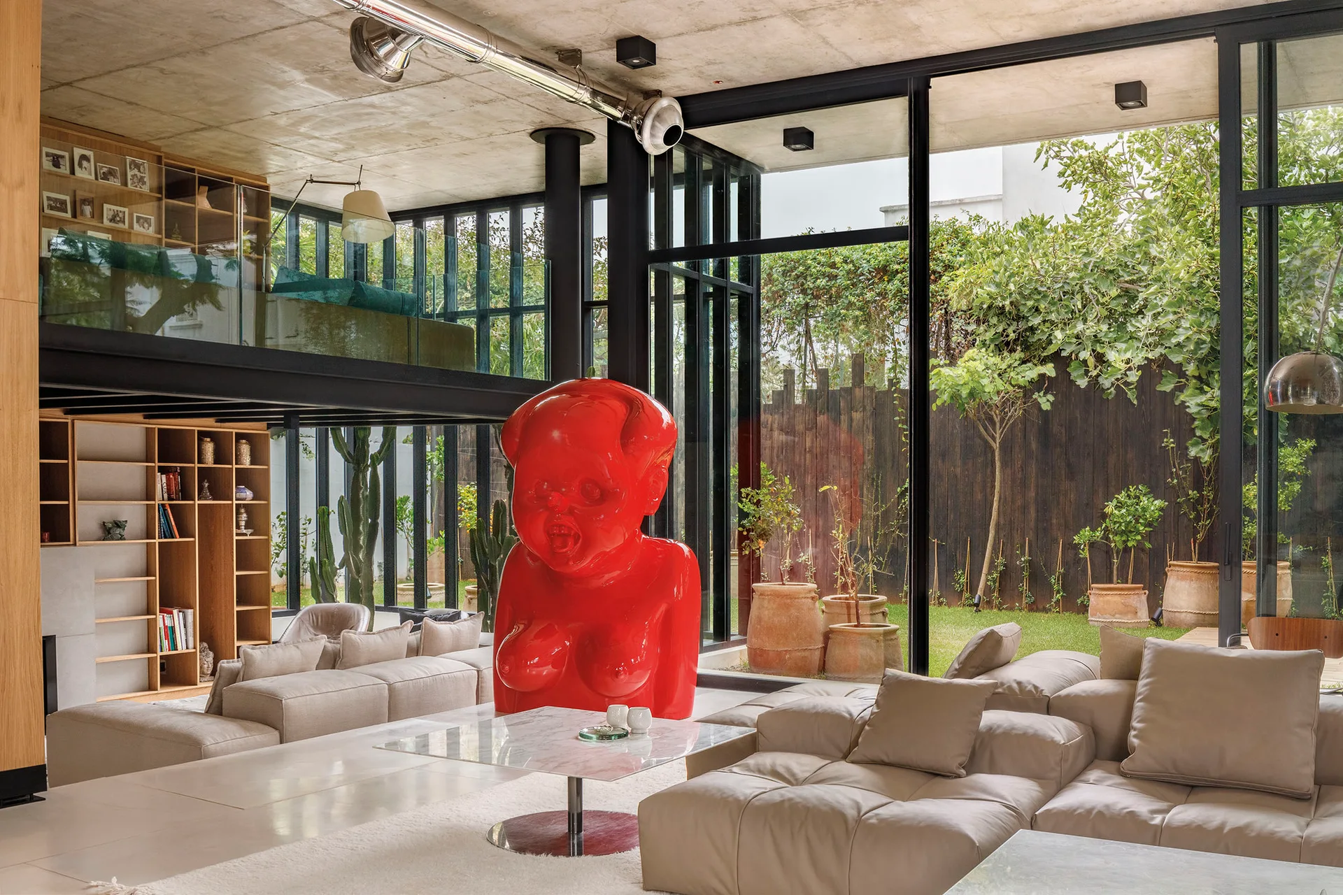 A spacious modern living room featuring a large glossy red sculpture, light beige modular sofas, a raw concrete ceiling, and floor-to-ceiling windows.