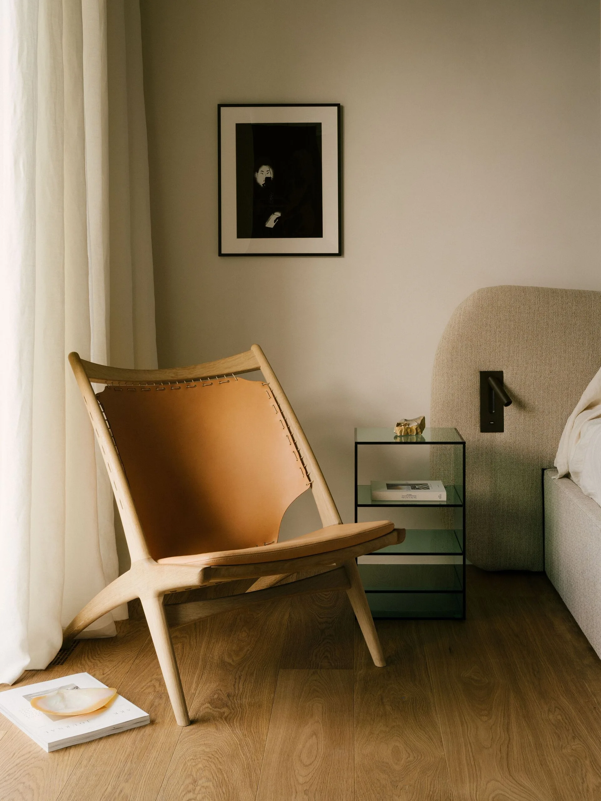 A tan leather sling chair sits in a bedroom corner next to a green glass bedside table, an upholstered headboard, and a framed black-and-white print.