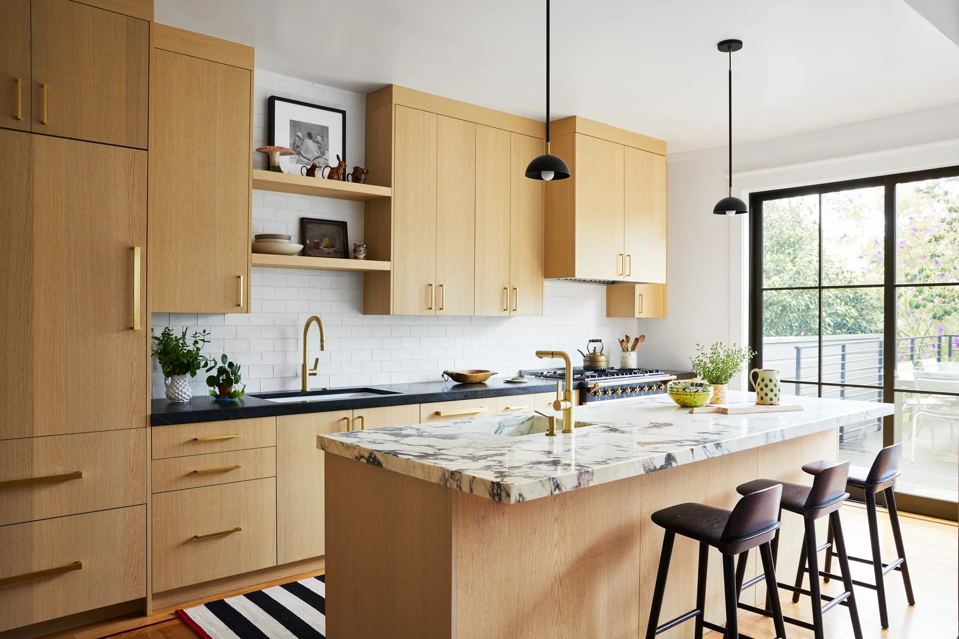 A contemporary kitchen featuring flat-front light wood cabinets, a white marble-topped island with black bar stools, and black dome pendant lights.